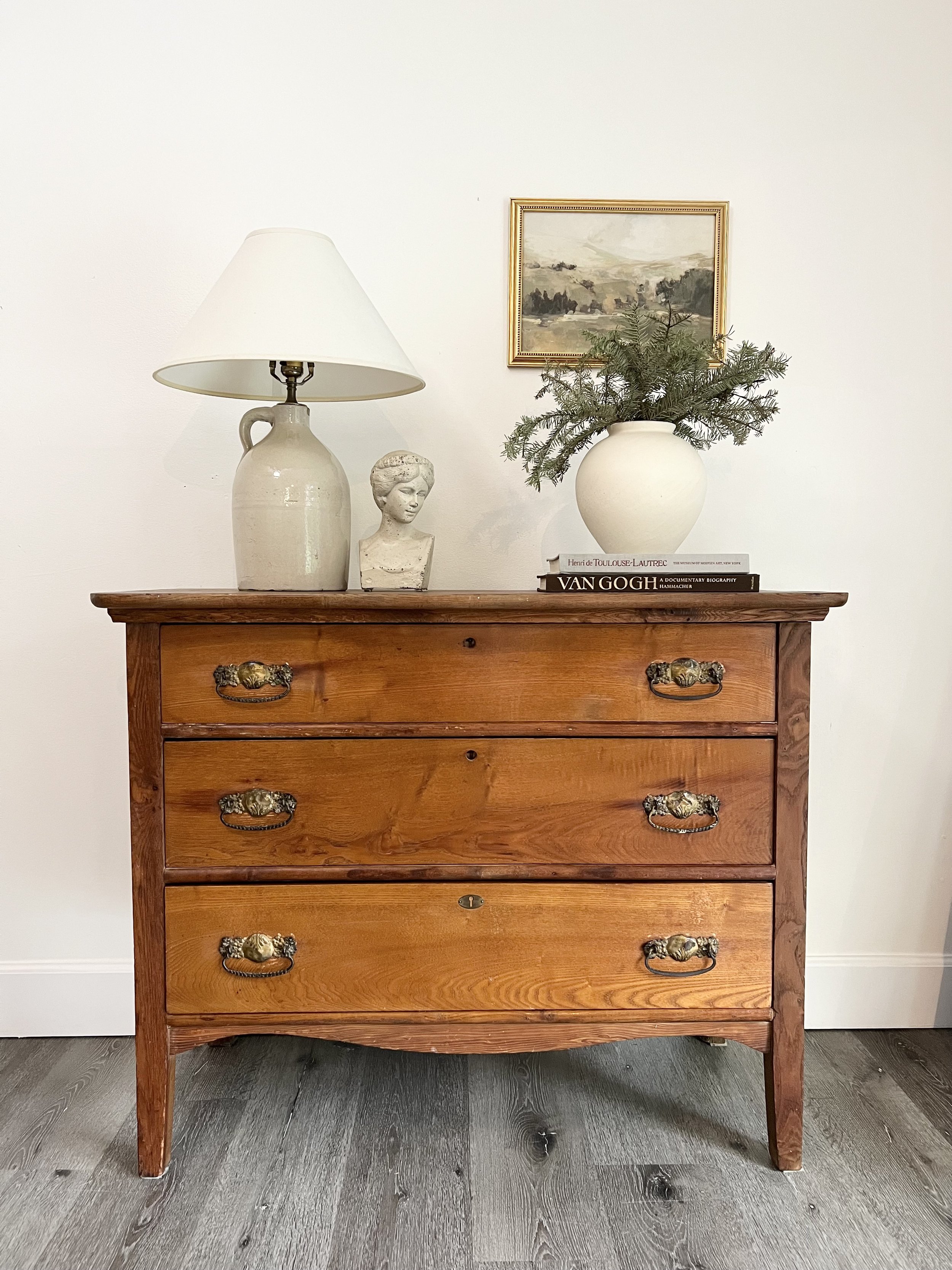 A wooden dresser with four drawers, brass handles, and a curved bottom edge, placed against a white wall. On top, there is a white ceramic table lamp with a beige shade, a small bust sculpture, a white vase with greenery, and three art and photograph