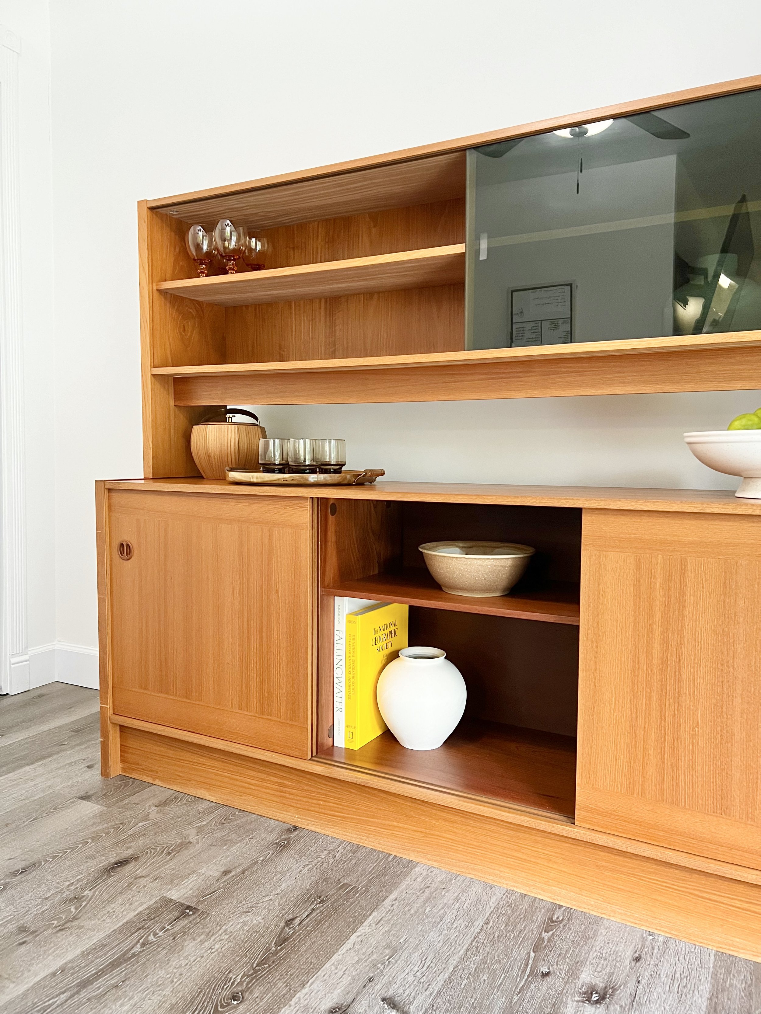 Wooden sideboard with glassware and decorative items, located on a light-colored wood floor, against a white wall.