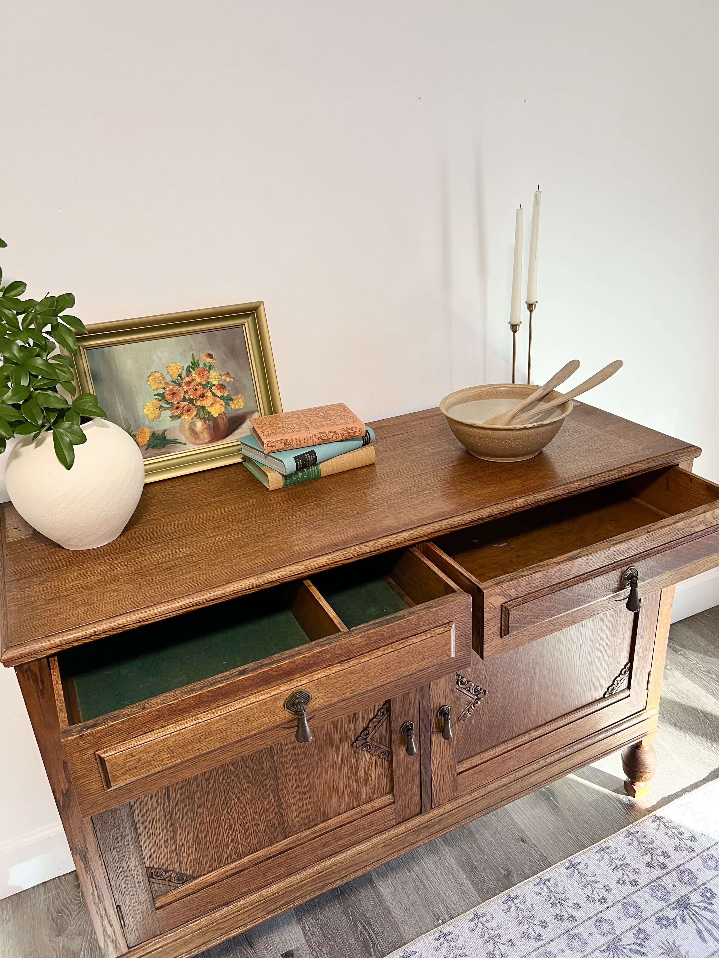 Vintage wooden sideboard with open drawers, decorative items on top including a framed painting of flowers, a potted plant, stacked books, a ceramic bowl with wooden spoons, and two tall candles, positioned against a white wall with a patterned rug o