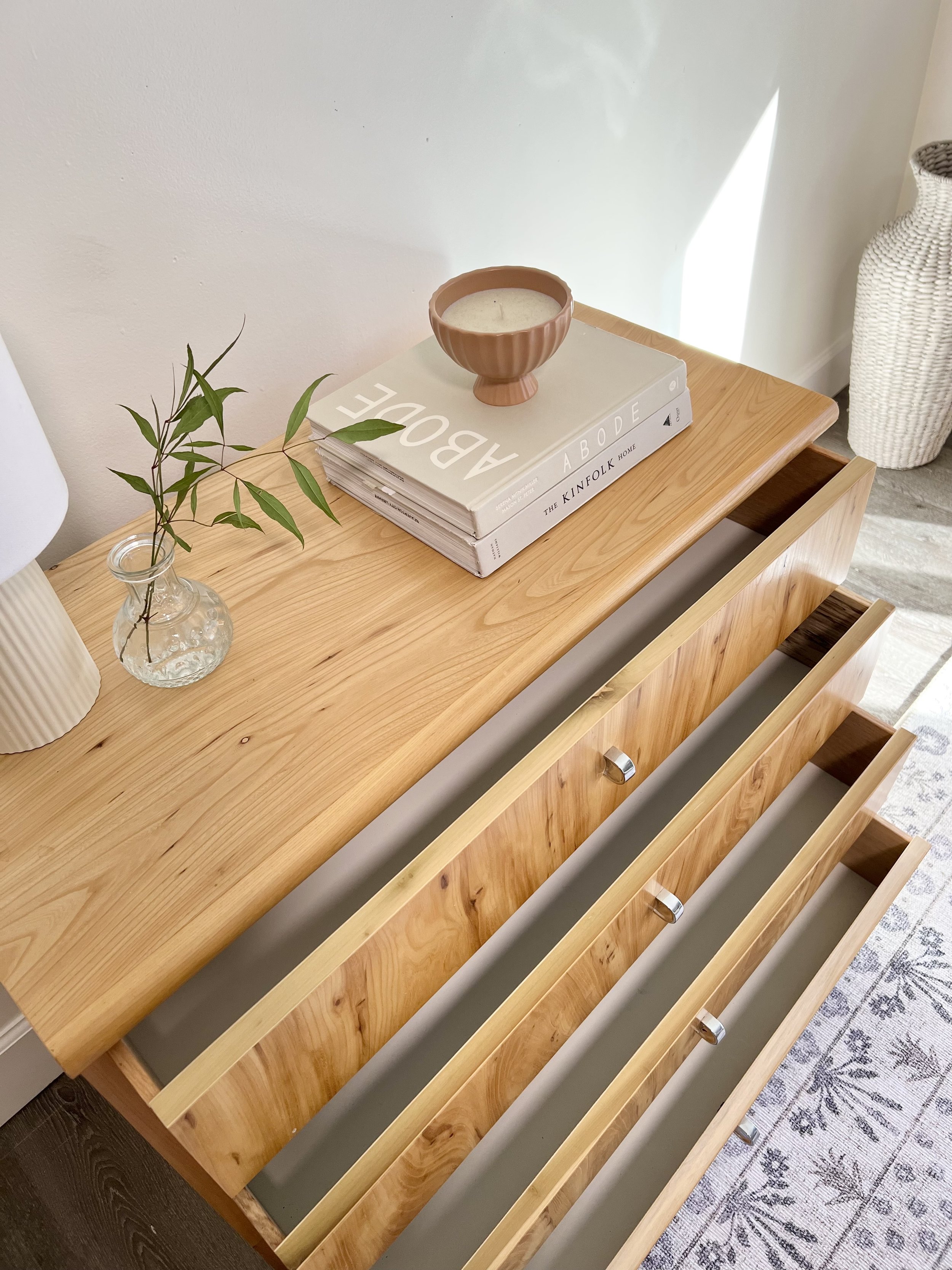 A wooden dresser with open drawers, a small glass vase with green leaves, a white ribbed lamp base, a pile of magazines or books, a pink ceramic bowl, and a white wicker basket in the background.