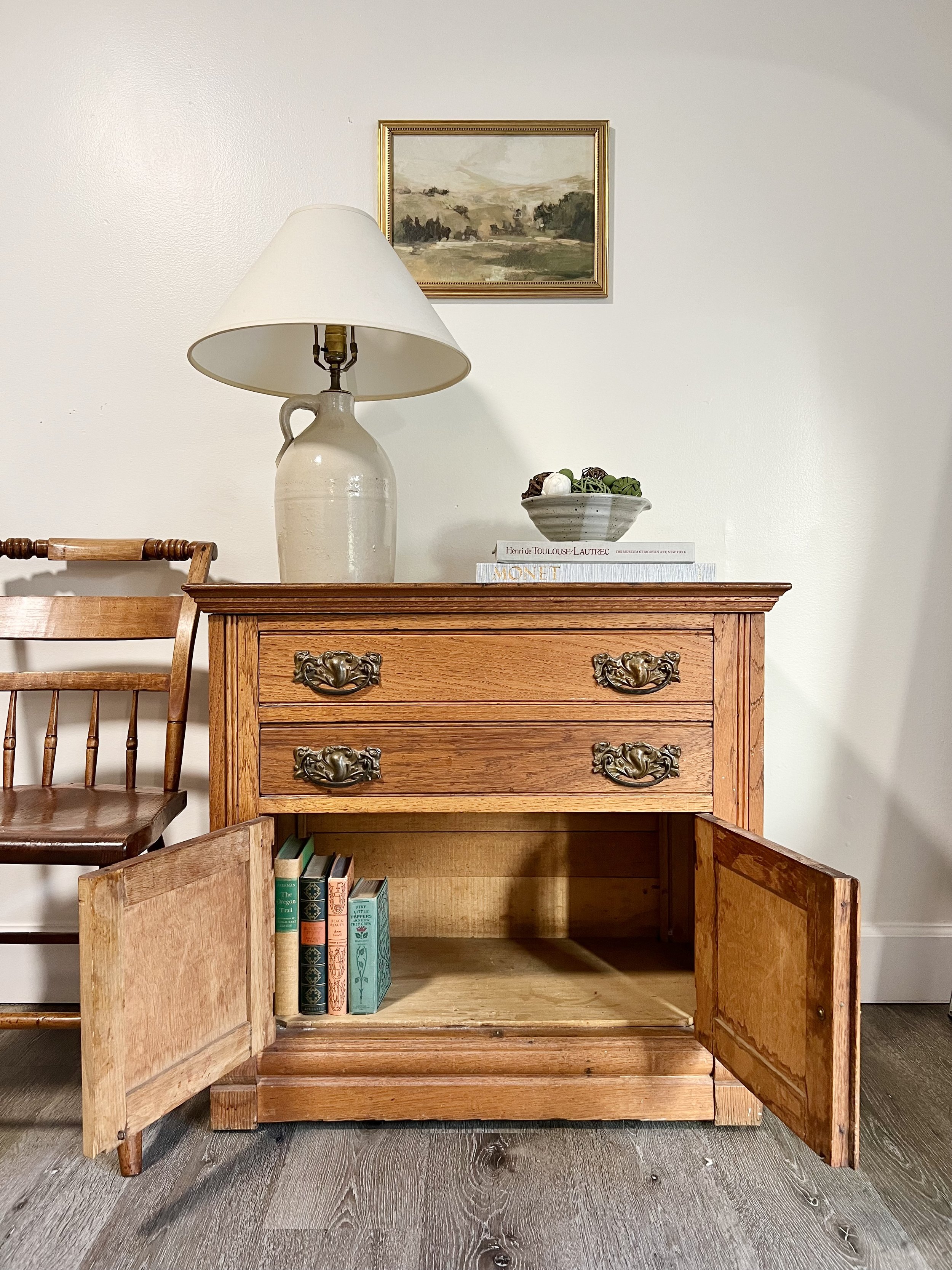 A wooden cabinet with open doors revealing a few books inside, topped with a ceramic lamp with a white shade, a bowl of decorative items, and a stack of art books. A framed landscape painting hangs on the wall above.