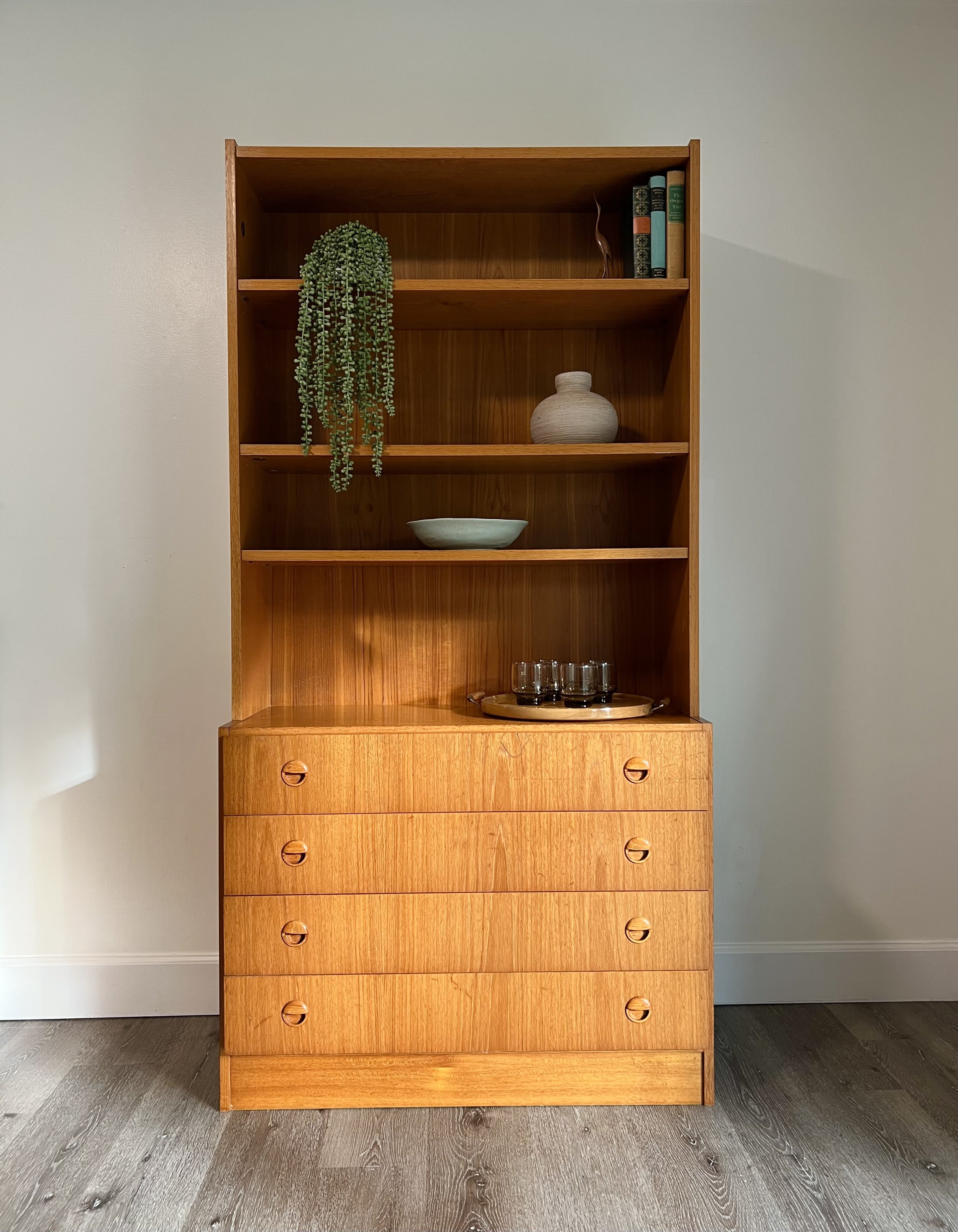 Wooden bookshelf with four drawers at the bottom, decorated with books, ceramics, and a plant, against a light gray wall.