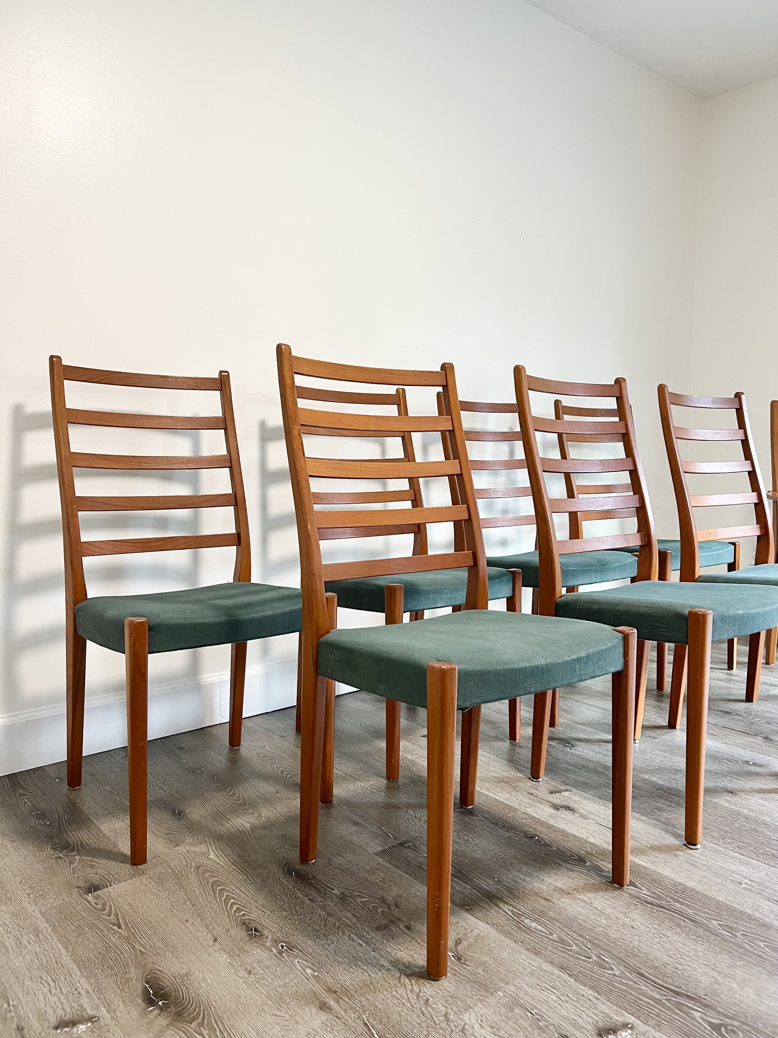 Six wooden chairs with green cushioned seats arranged in a row against a plain white wall and wood-textured floor.