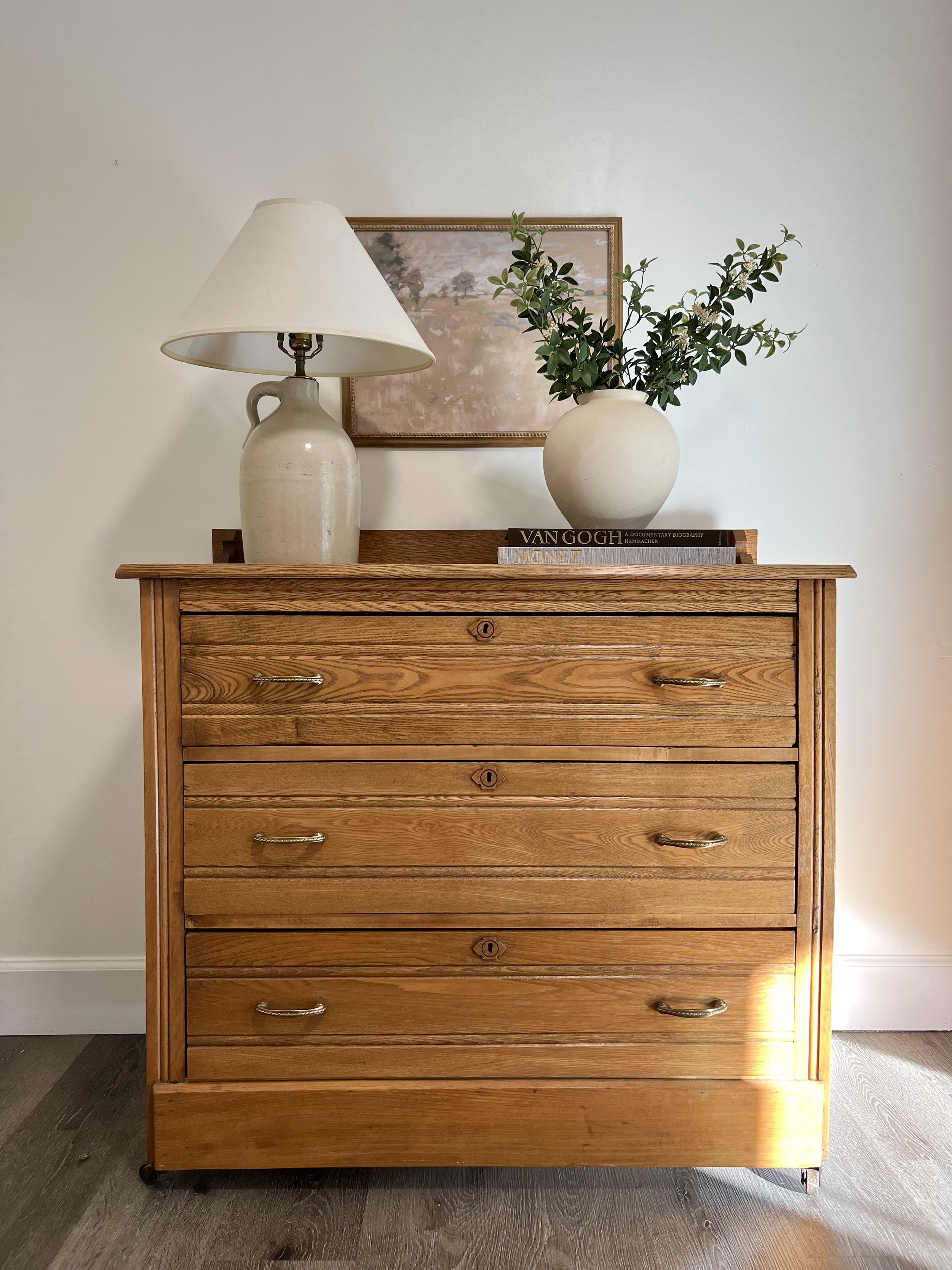 A wooden dresser with five drawers against a white wall, topped with a table lamp, a vase with greenery, and two books, with a framed landscape painting hanging above.