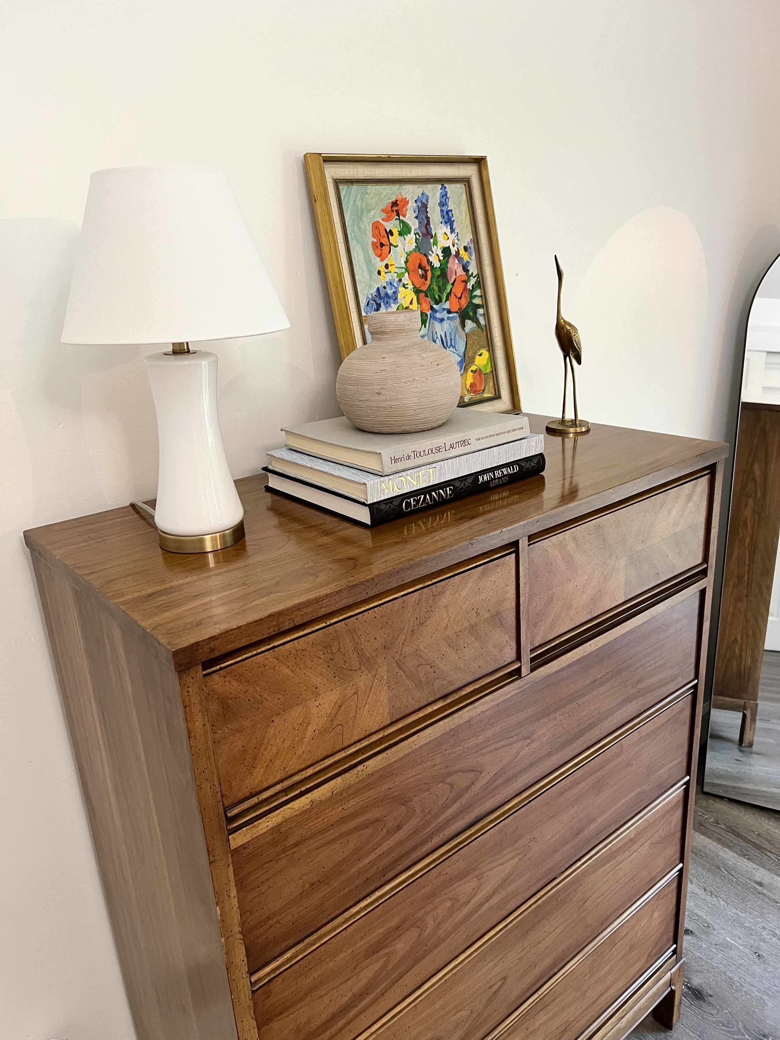 A wooden dresser with a white lamp, framed colorful floral artwork, a beige vase, a stack of art and design books, and a gold heron figurine on top.