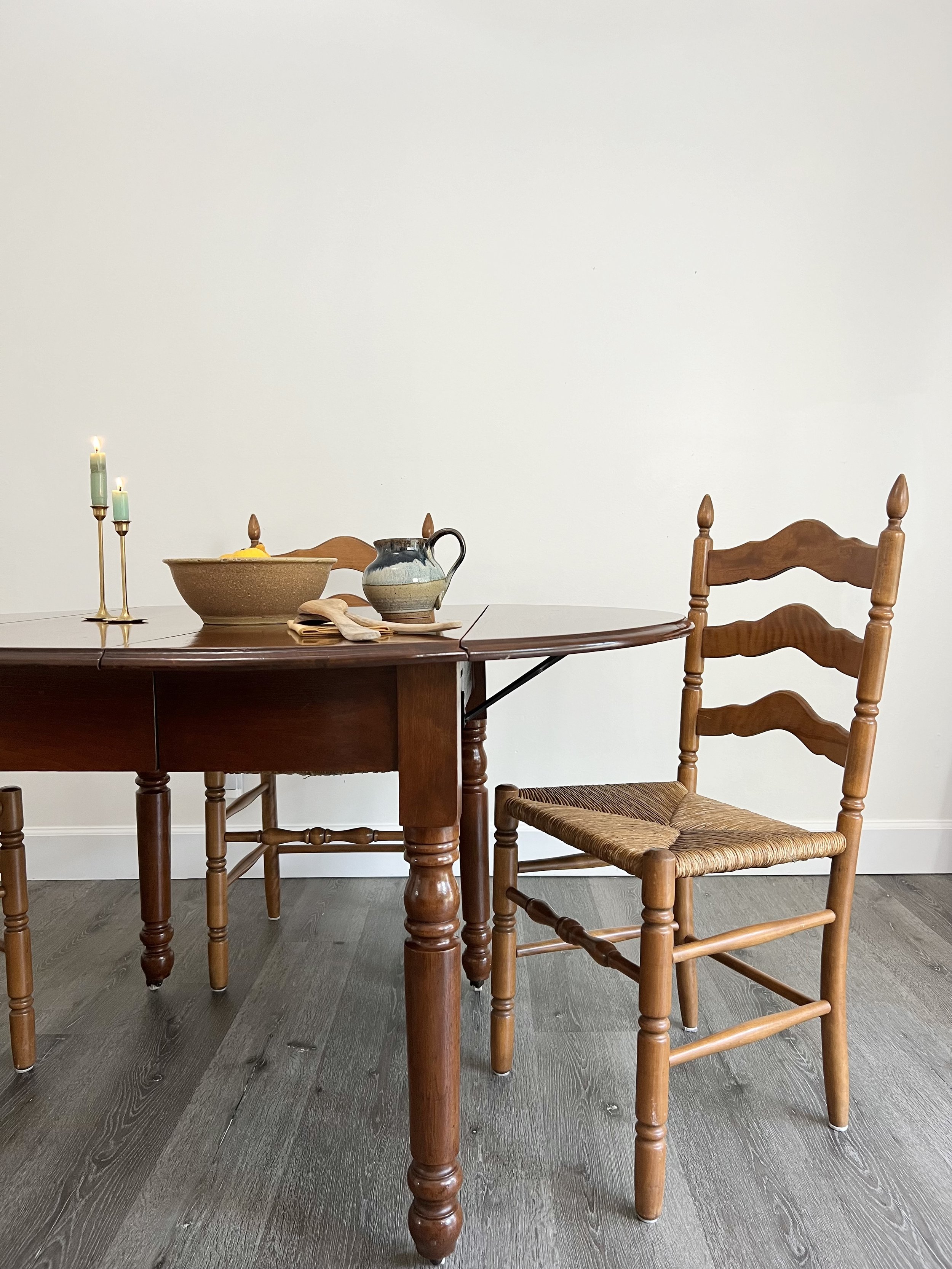 A wooden dining table with a chair beside it, decorated with a candle holder, a bowl, a pitcher, and some utensils against a plain white wall.