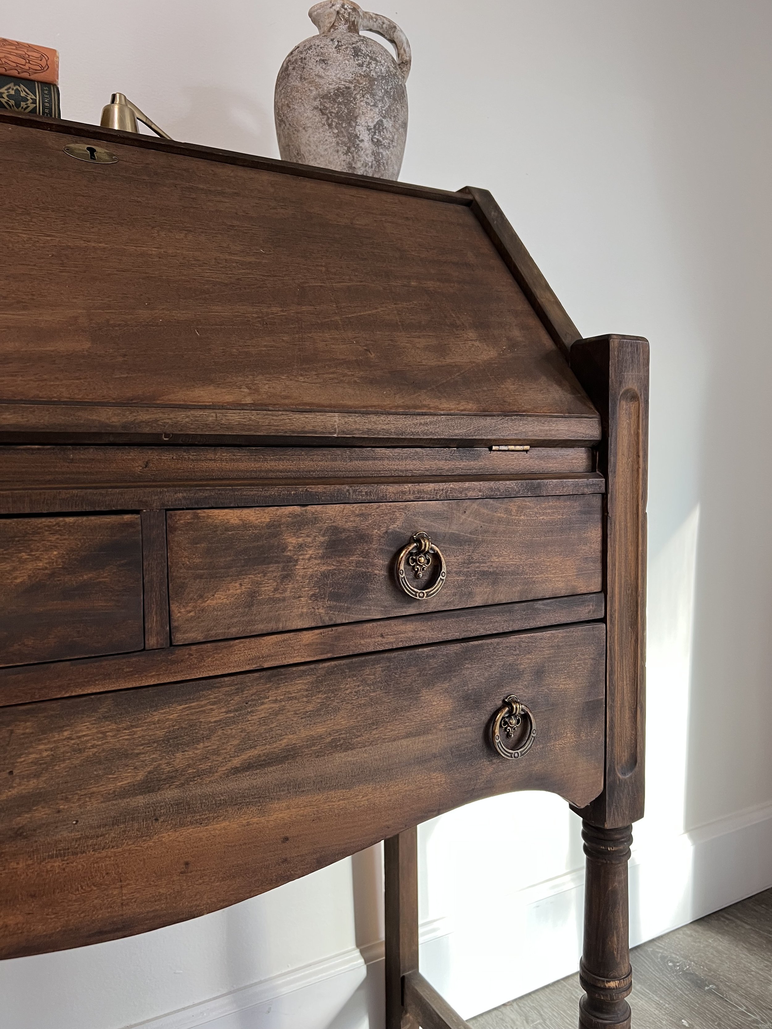 A vintage wooden writing desk with brass ring pulls on drawers, a rustic ceramic pitcher on top, and a brass desk lamp on the side, set against a white wall.