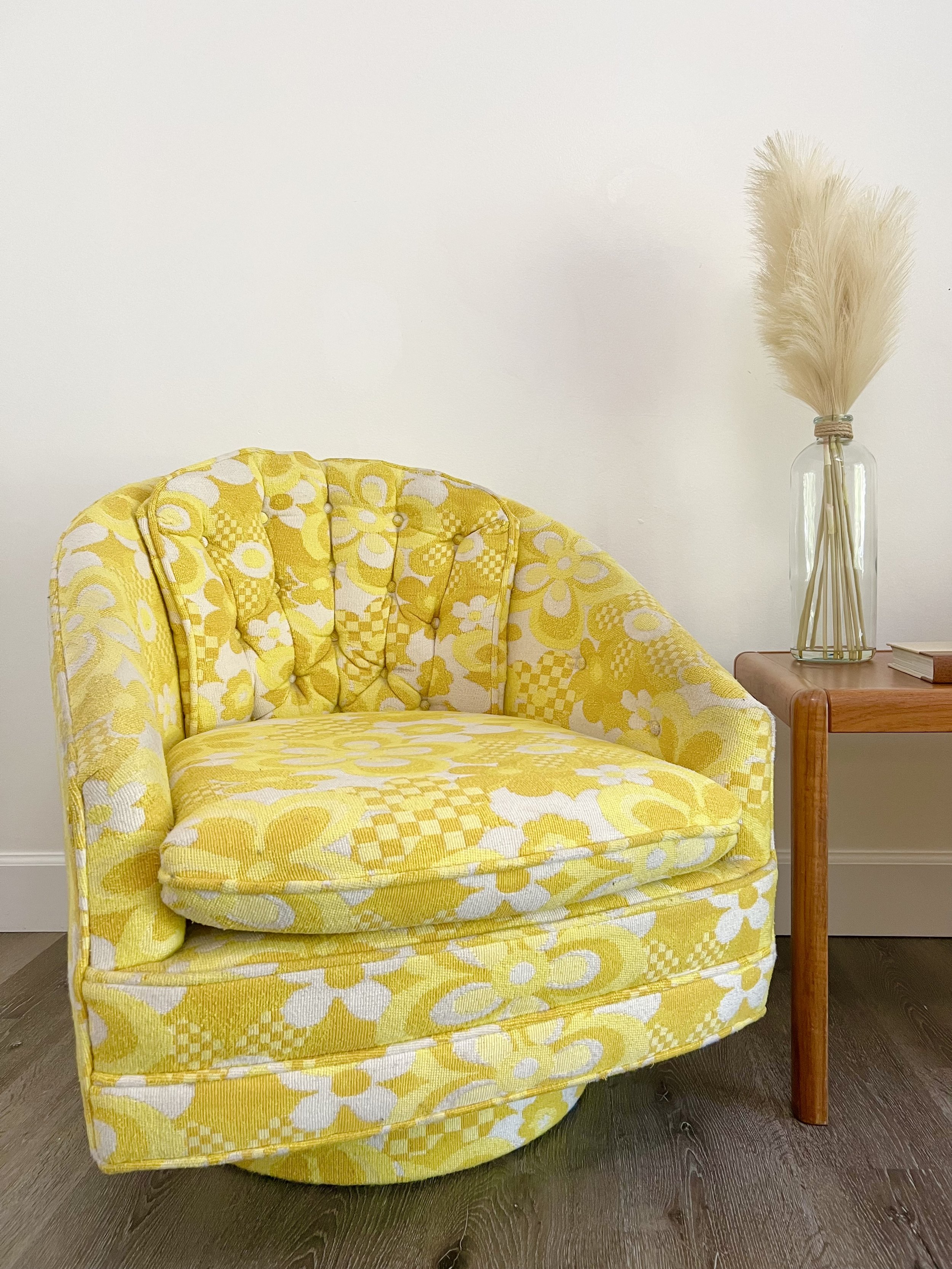 Yellow floral-patterned upholstered armchair next to a wooden side table with a glass vase holding dried pampas grass.