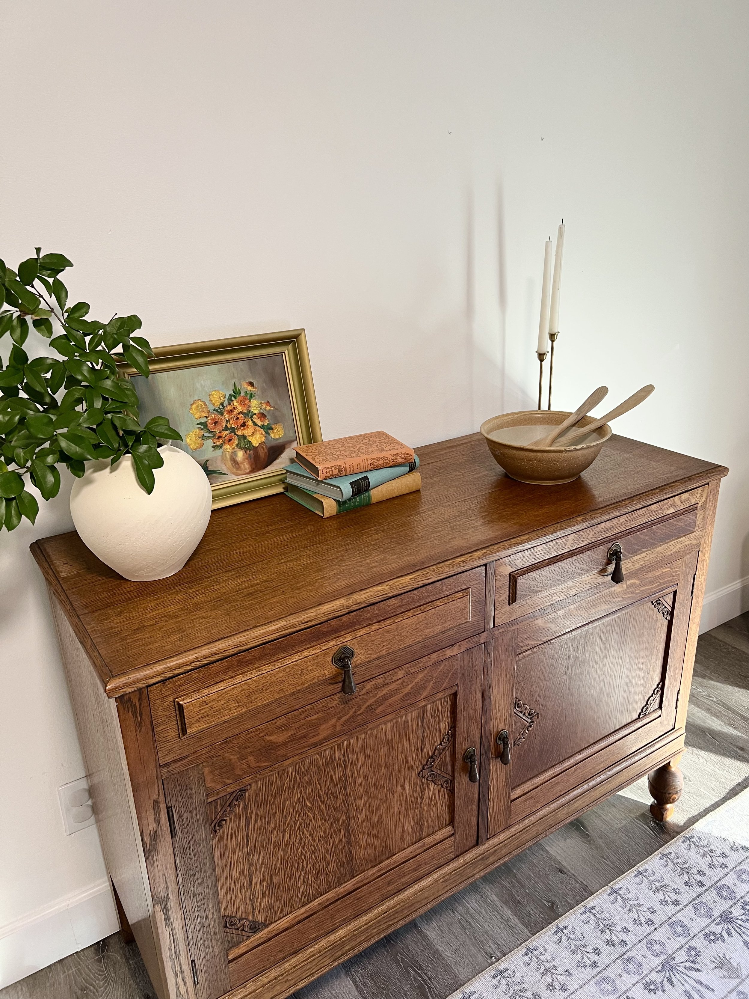A wooden sideboard with decorative carvings and round wooden feet. On top, there is a potted plant with green leaves, a framed floral painting, three stacked books, a brown bowl with two wooden spoons, and two white candles in brass holders.