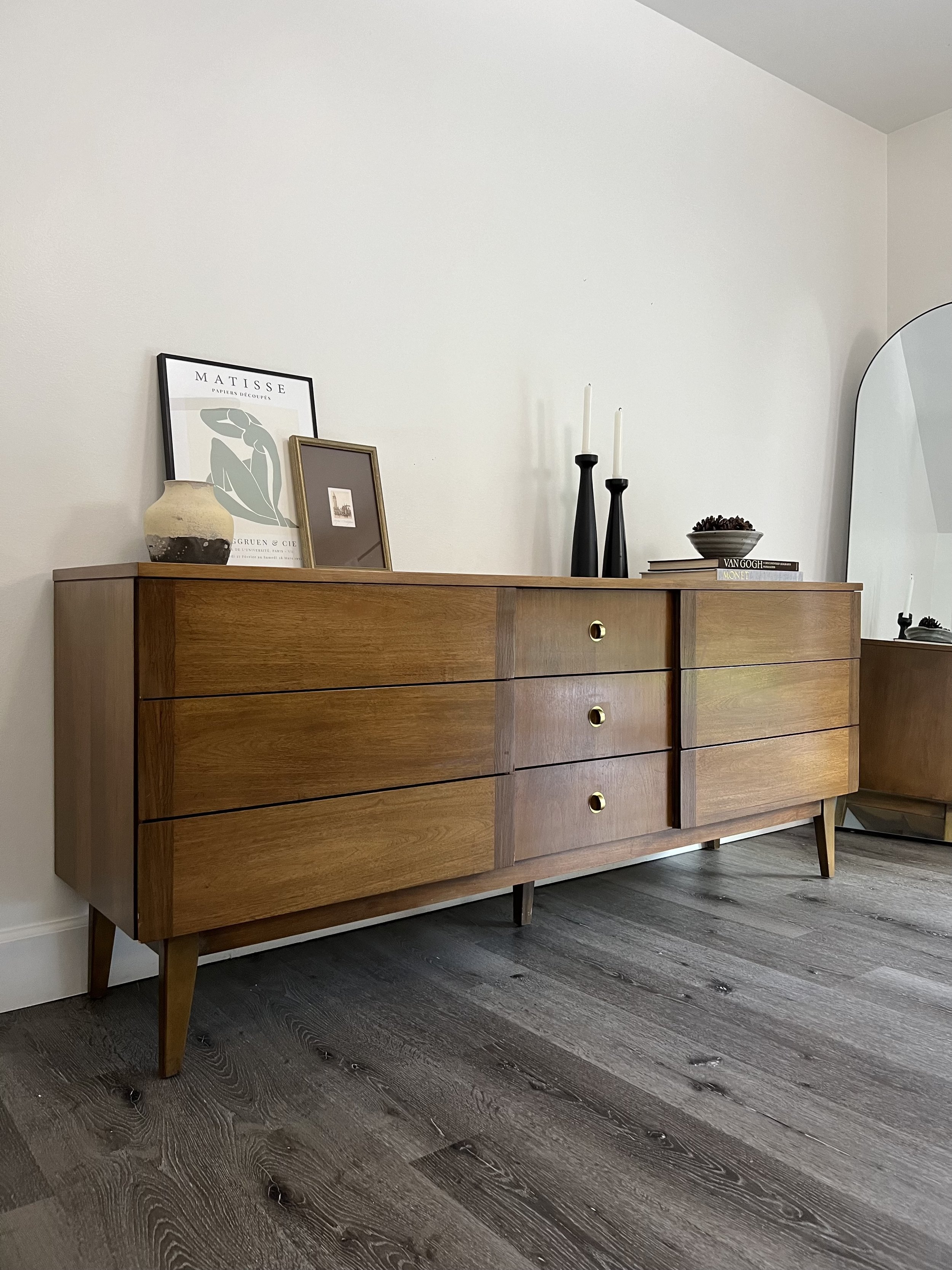 Mid-century modern wooden sideboard with four drawers and three cabinet doors, decorated with framed artwork, black candlesticks, books, and a ceramic vase, against a neutral wall.