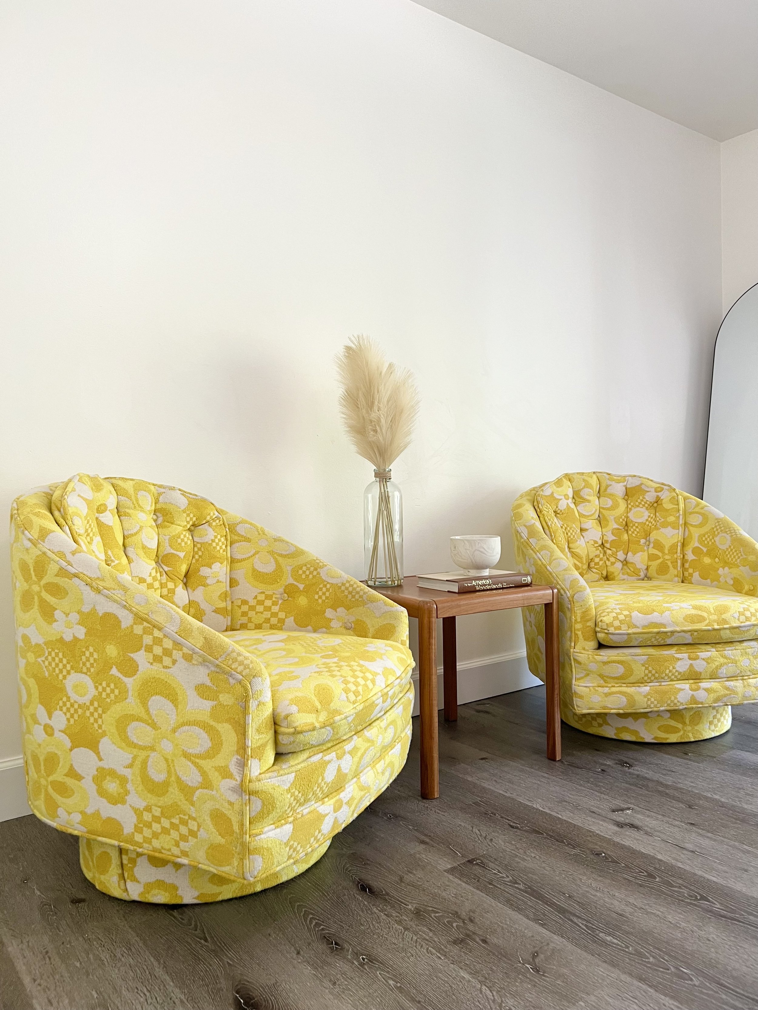 Two yellow floral-patterned armchairs with tufted backs flank a small wooden side table with a glass vase of pampas grass, a white bowl, and books, set against a white wall and dark wood floor.