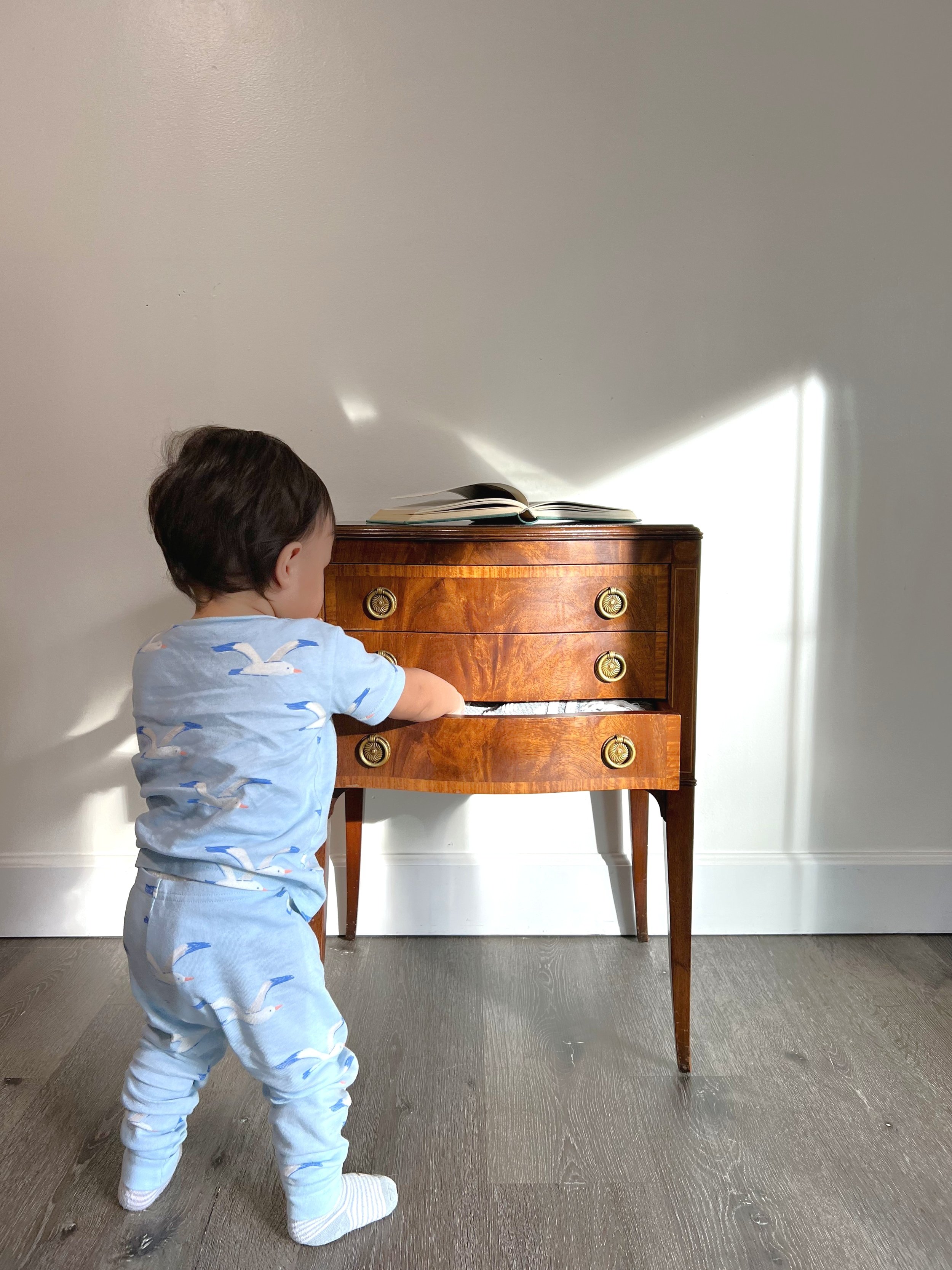 A young child in light blue pajamas with seagull patterns reaching into an open drawer of a small vintage wooden dresser. An open book sits on top of the dresser, which is against a plain light-colored wall with sunlight casting patterns on it.