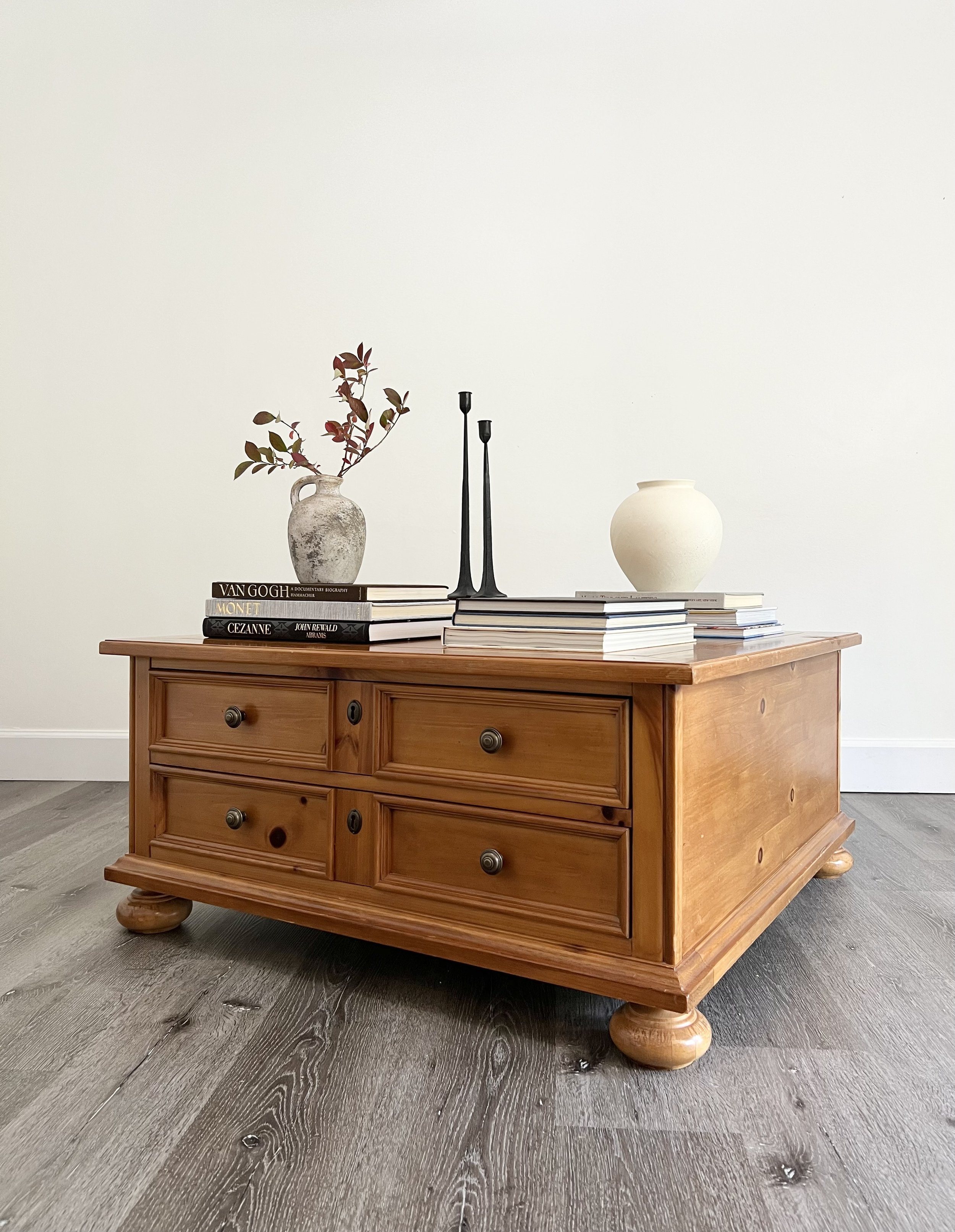 A wooden coffee table with four drawers and rounded feet, decorated with vases, books, and candle holders, set against a plain wall and dark wooden floor.