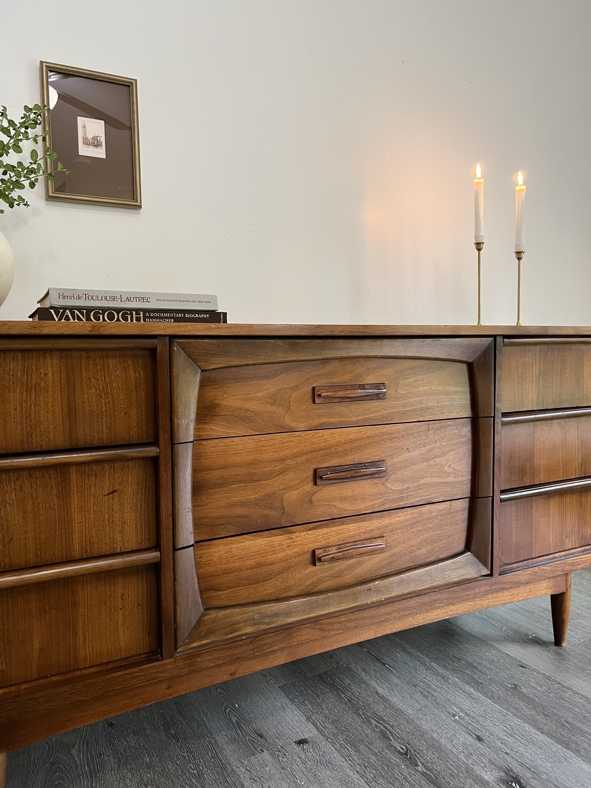 Mid-century wooden sideboard with curved drawers in a room interior, decorated with books, framed art, a plant, and lit candles.