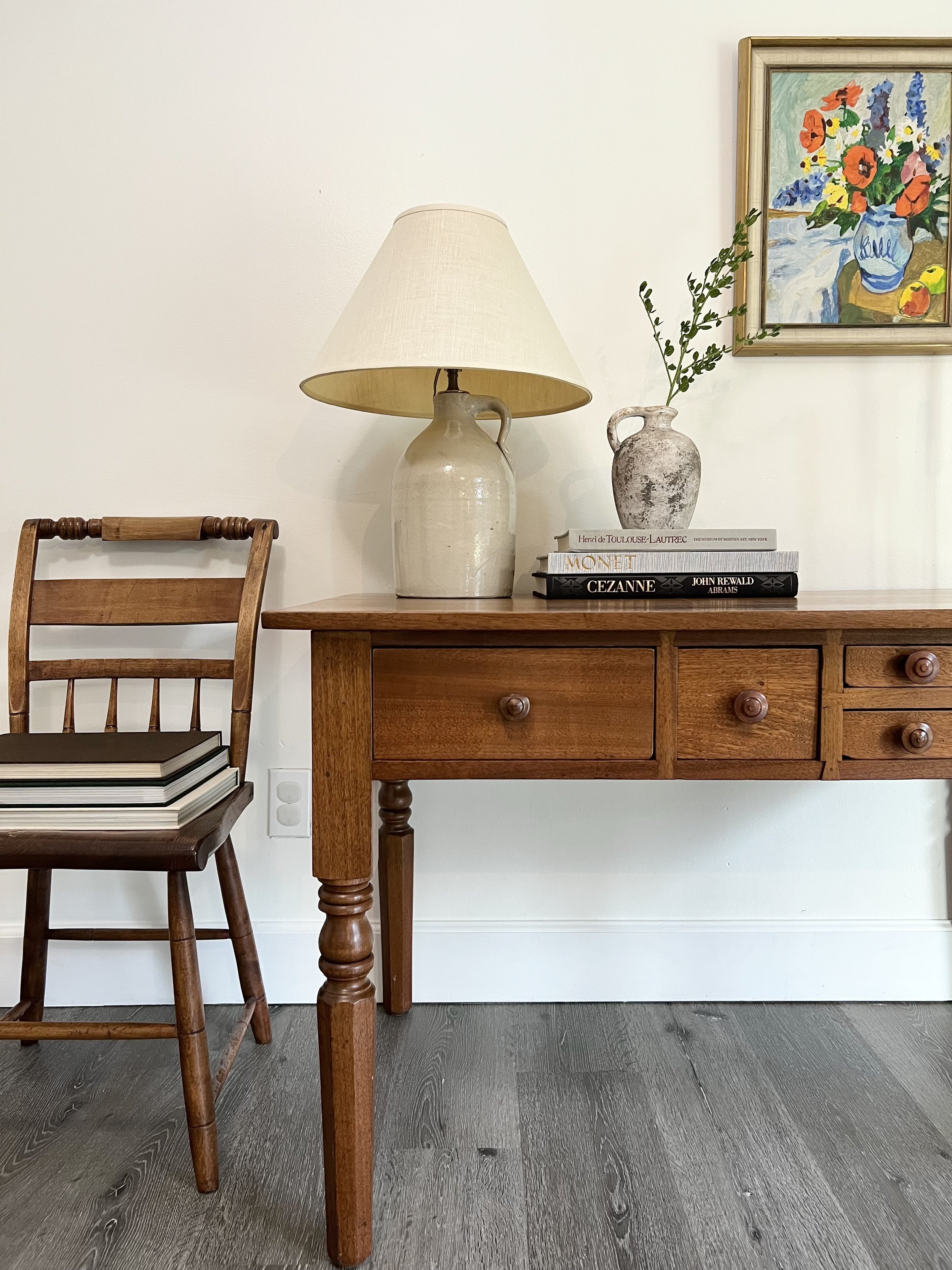 A wooden chair with stacked books on its seat, a wooden desk with a lamp and decorative vases, and a framed colorful floral painting on a white wall.