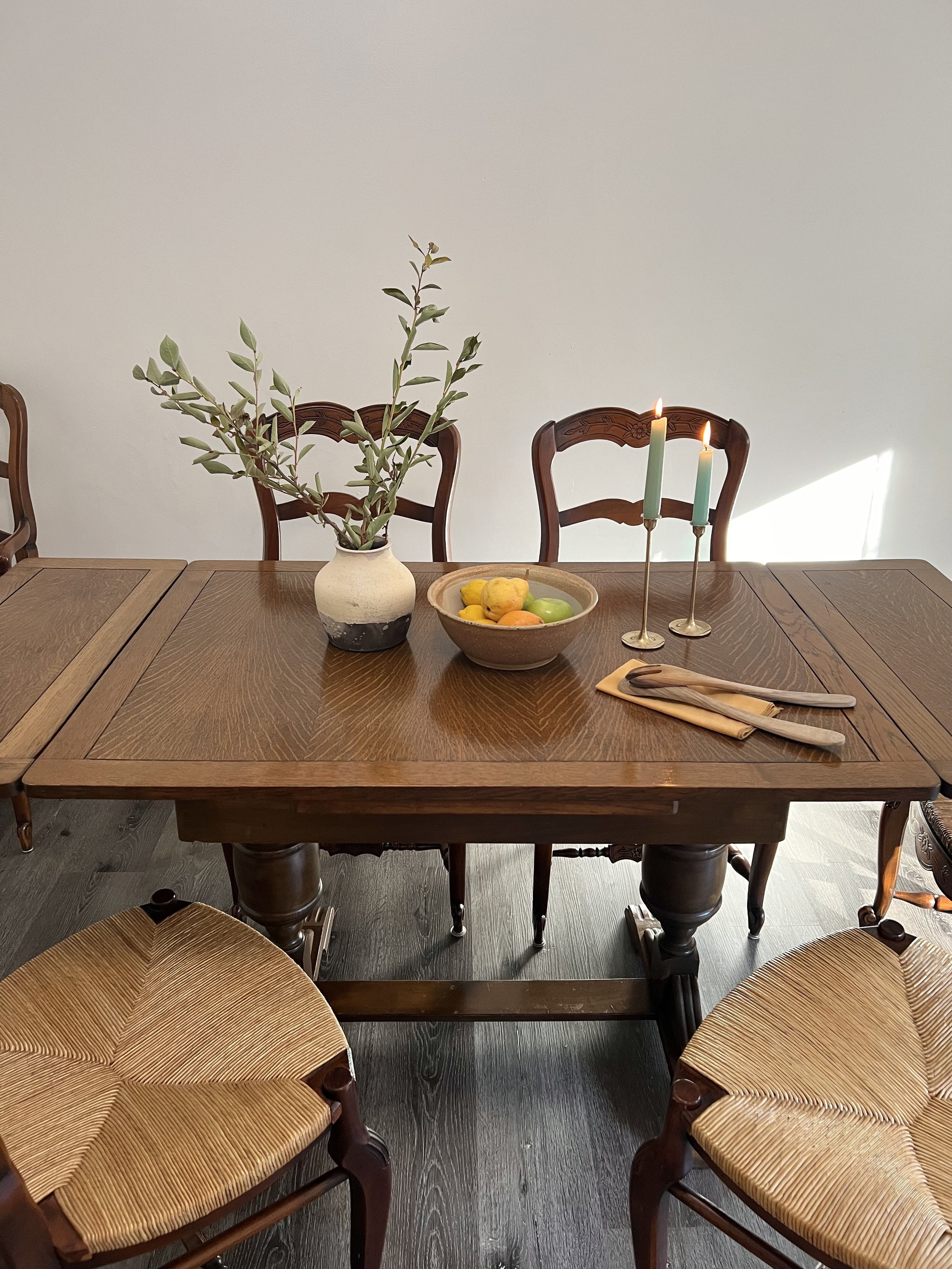 A wooden dining table with a vase of green foliage, a bowl of colorful fruit, two lit candles, and folded napkins and utensils, surrounded by four wooden chairs with woven seats.