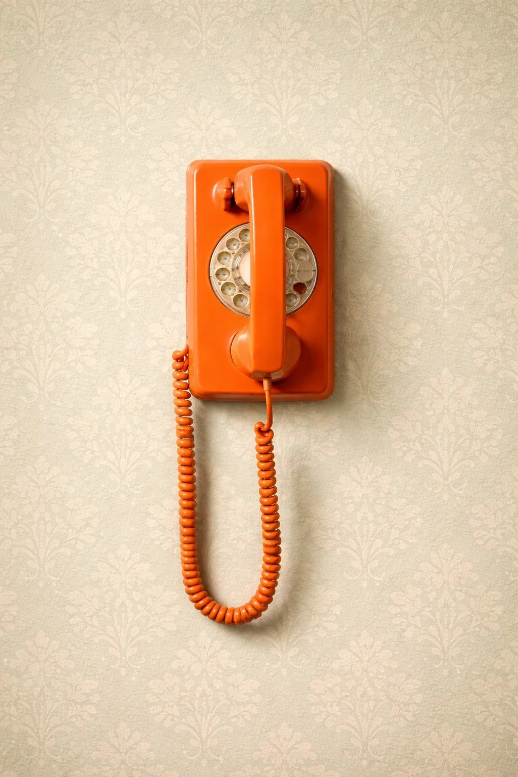 Orange vintage rotary telephone on a beige patterned wallpaper wall.
