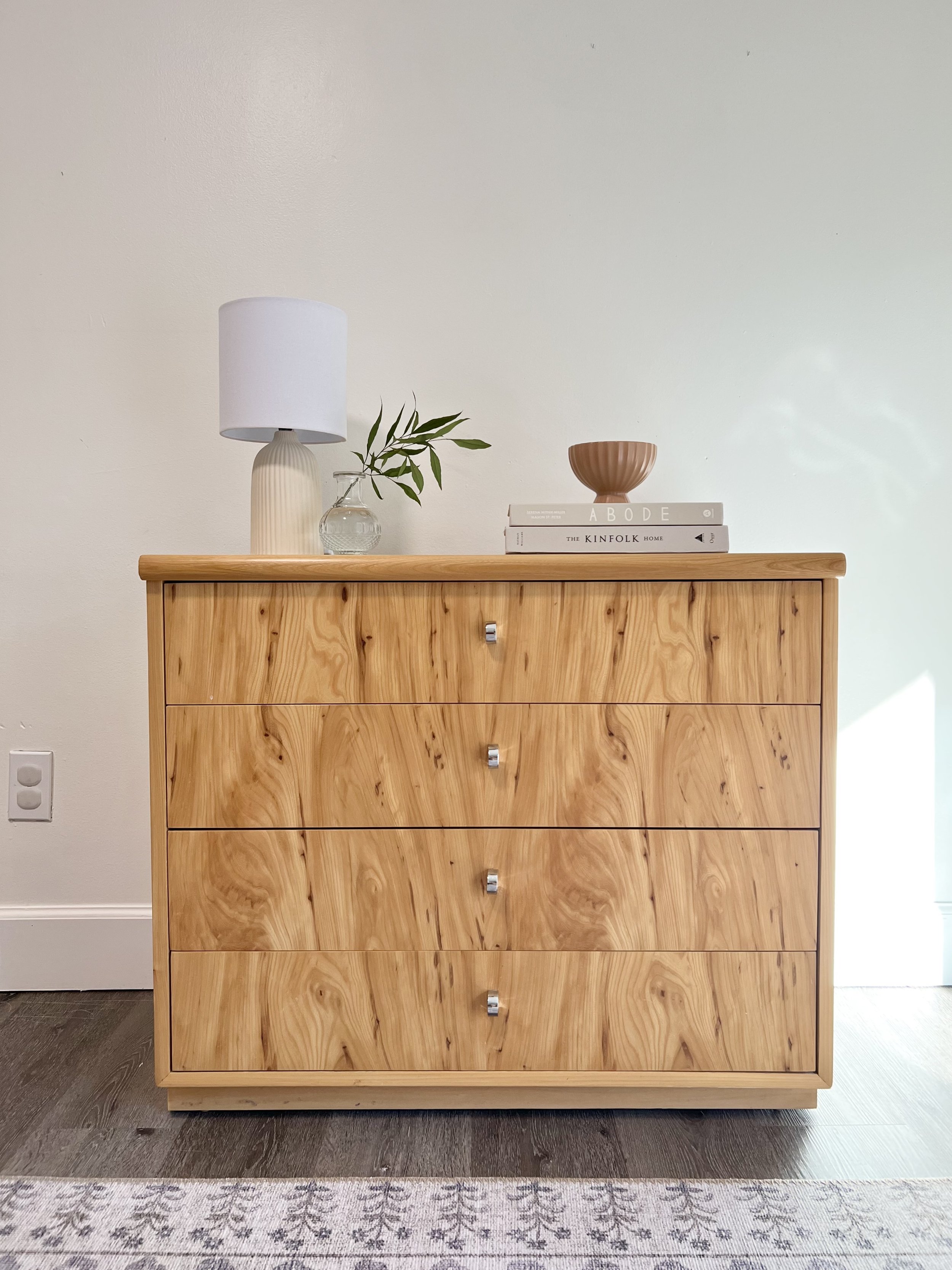 Wooden dresser with five drawers, topped with a white lamp, a glass vase with green leaves, a stack of three books, and a decorative bowl, against a white wall.