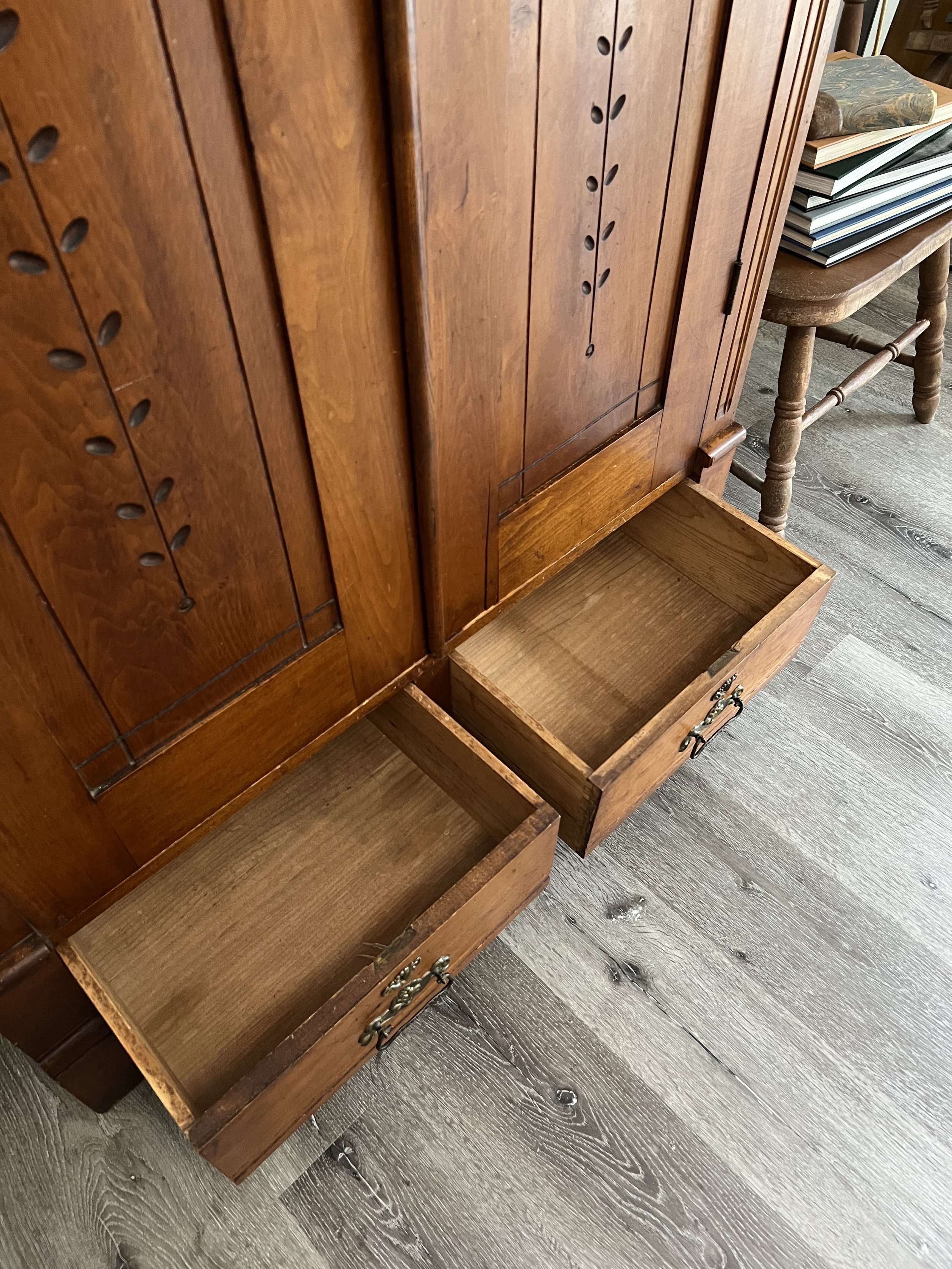 Open wooden drawers of a vintage wooden cabinet, with a pile of books resting on a chair beside it, on a wood floor.