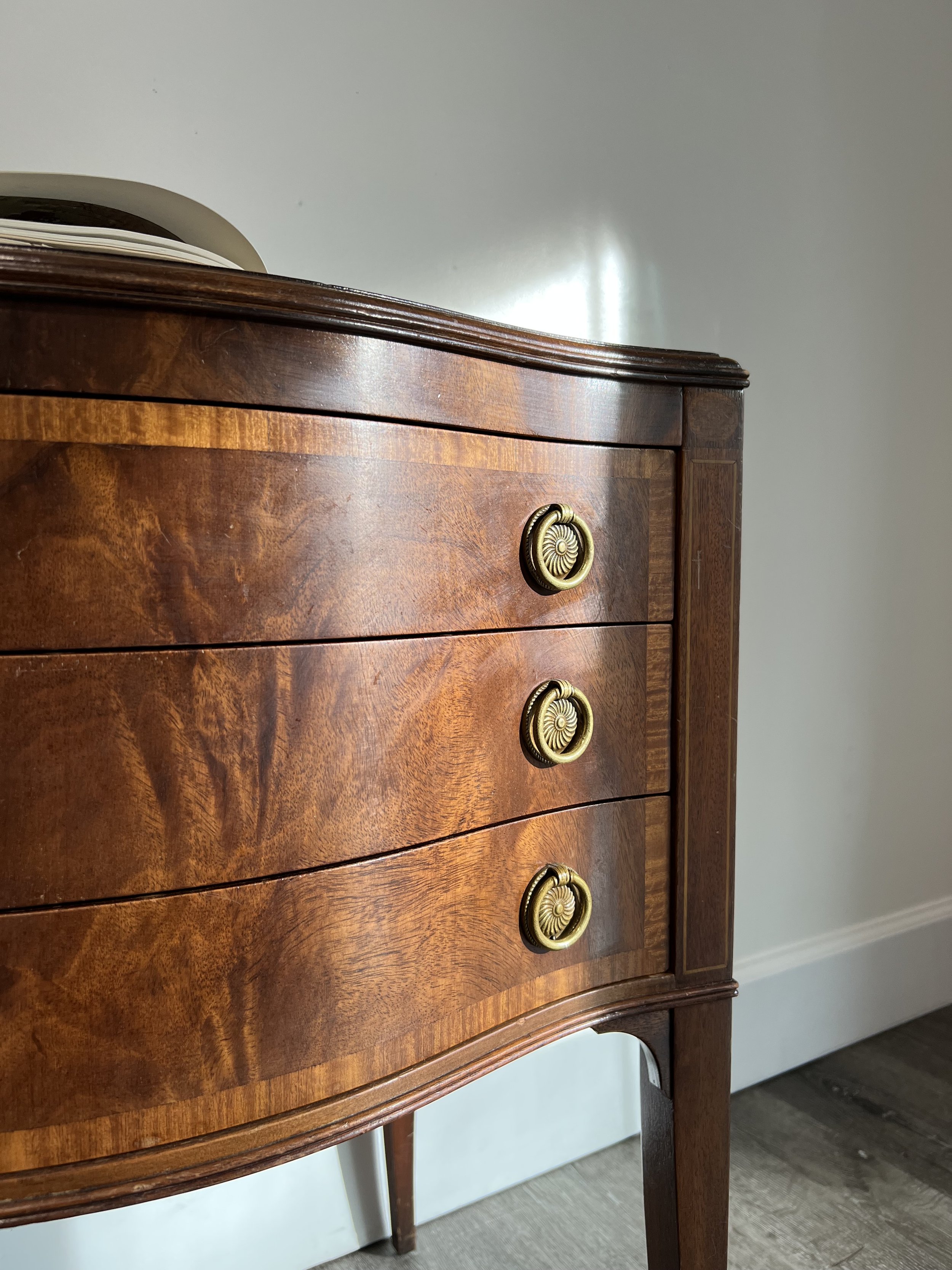 A vintage wooden dresser with three drawers, brass drawer pulls, and curved edges, standing on four tapered legs against a plain wall.