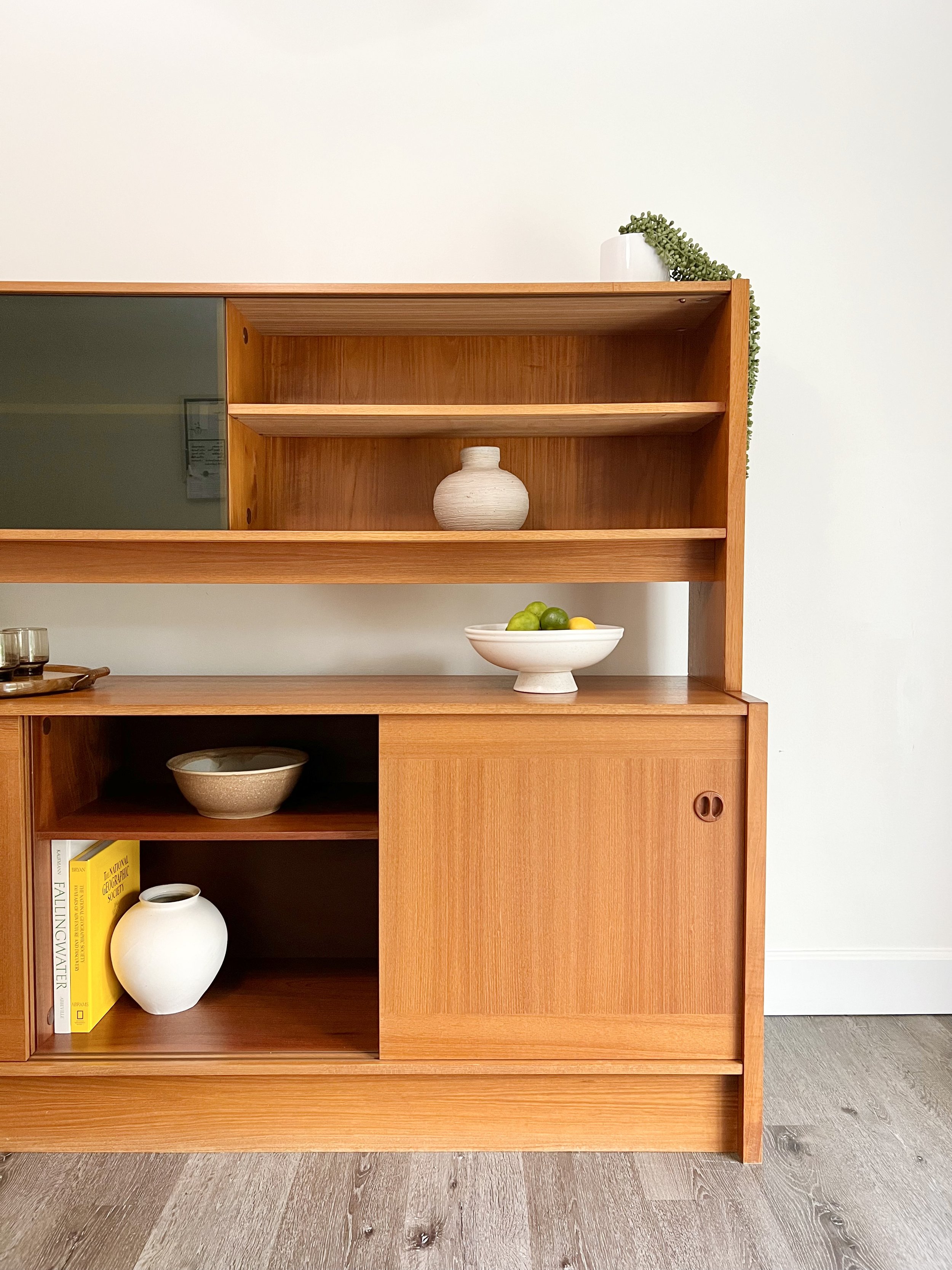 A mid-century modern wooden shelf with decorative vases, bowls, and books against a white wall and on a wooden floor.