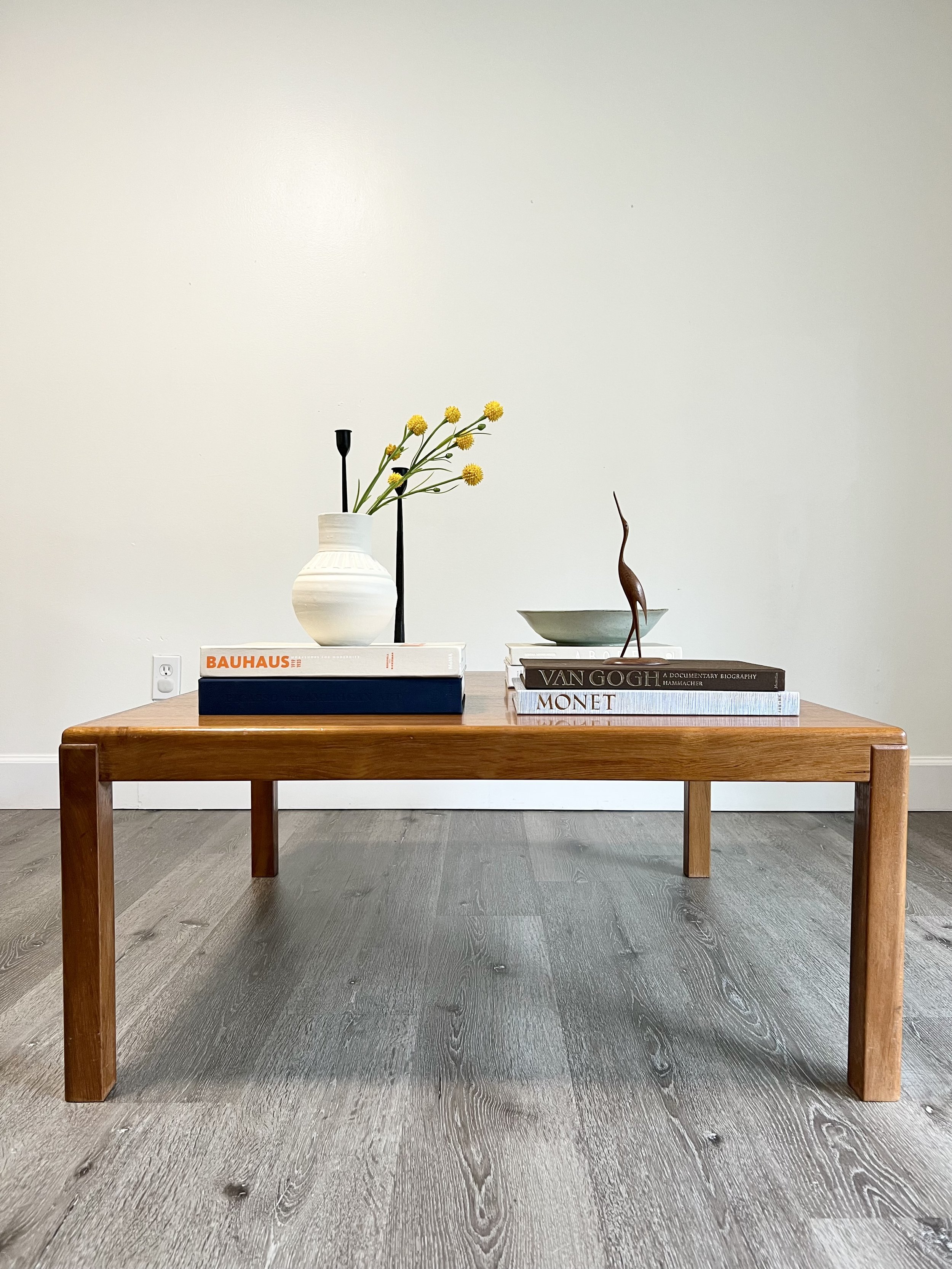 A wooden table with decorative items including a white vase with yellow flowers, a small sculpture, and stacks of art and design books against a plain white wall.