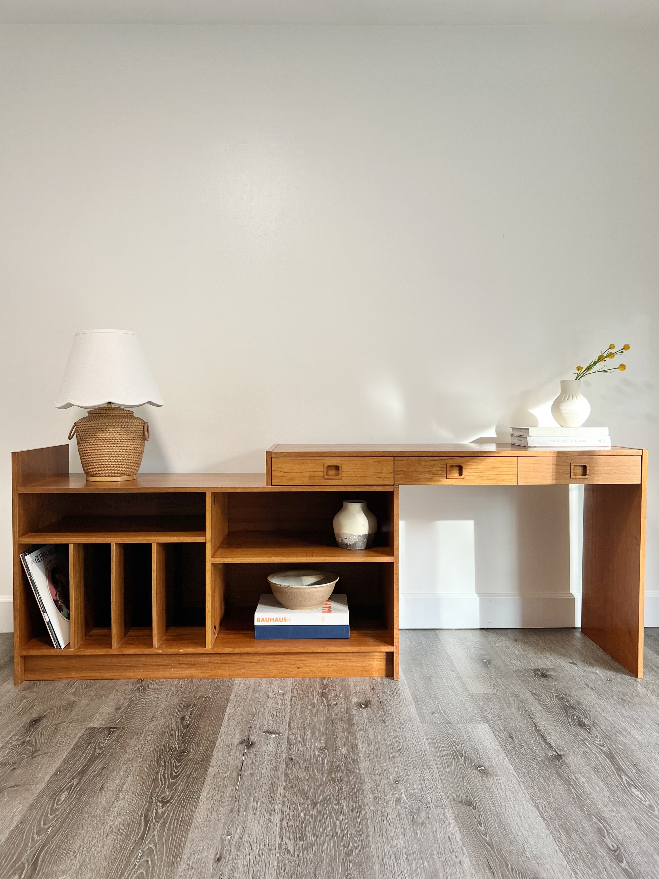 Wooden sideboard with decorative items on top, including a table lamp, vases, and books, against a white wall and wooden floor.