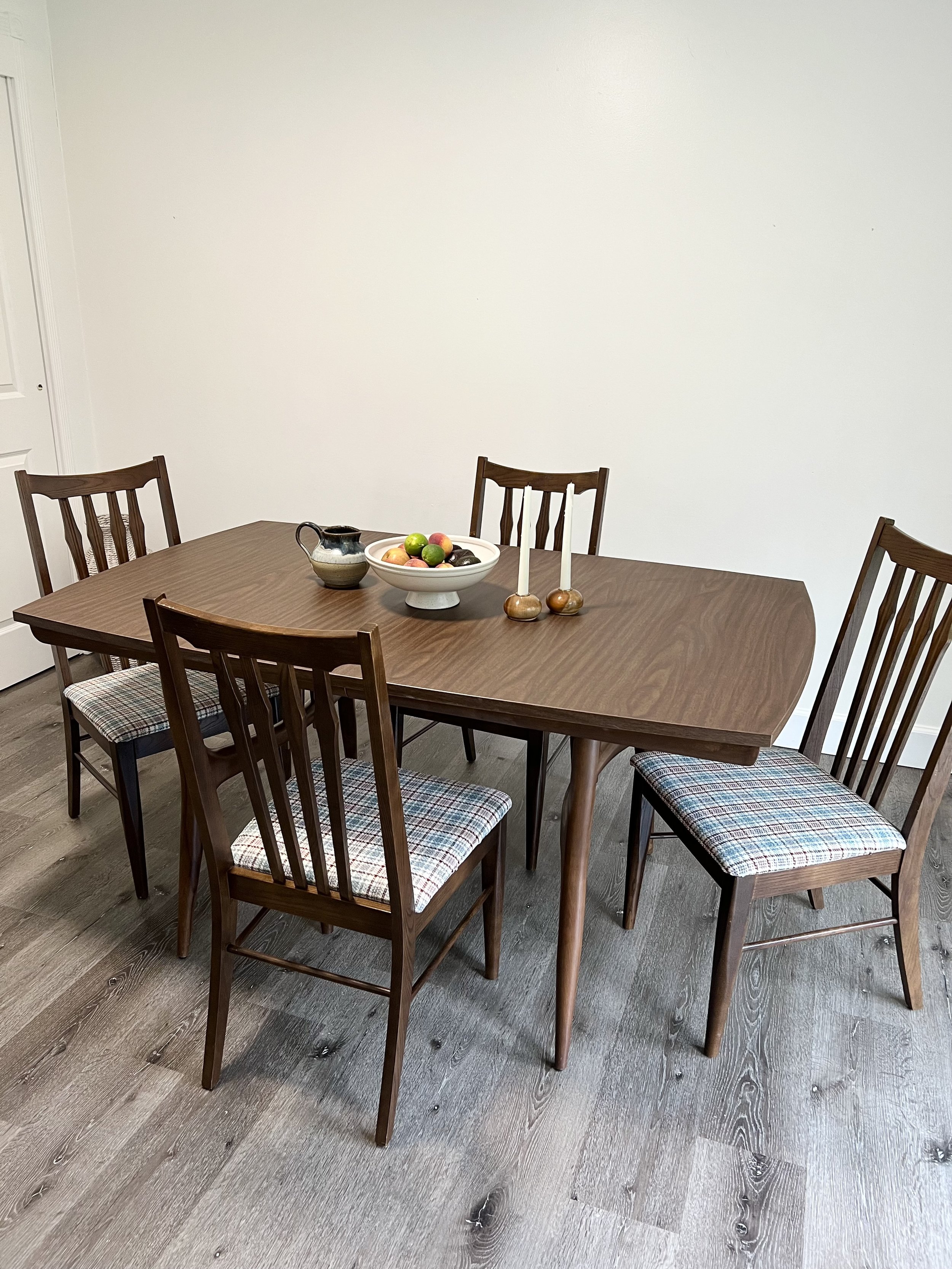 A dining table with five wooden chairs, a bowl of apples, two candles in candle holders, and a ceramic pitcher, all placed on a wooden floor against a plain white wall.