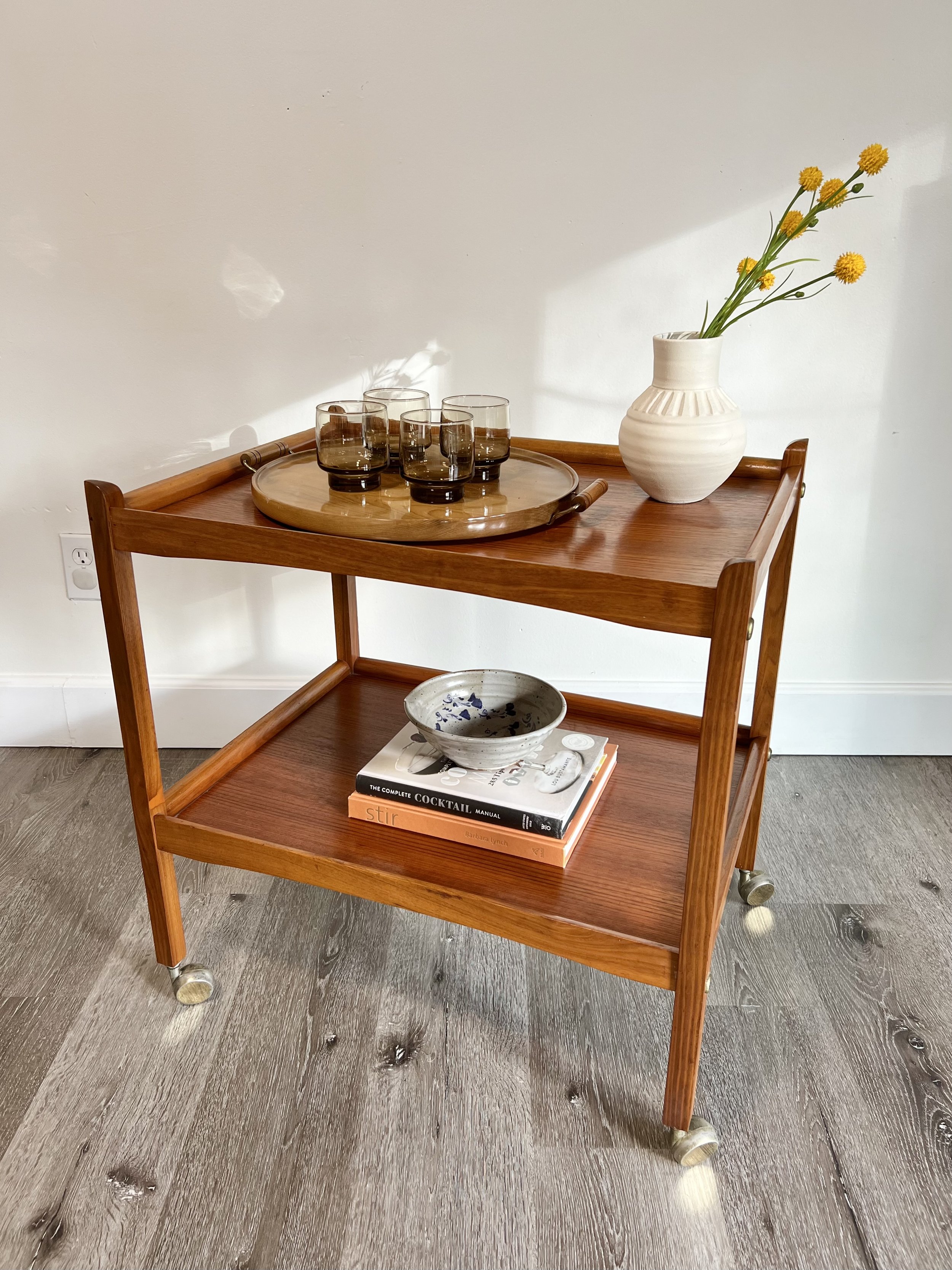 A wooden two-tier serving cart with glassware, a ceramic bowl on books, and a white vase with yellow flowers, set against a plain wall.