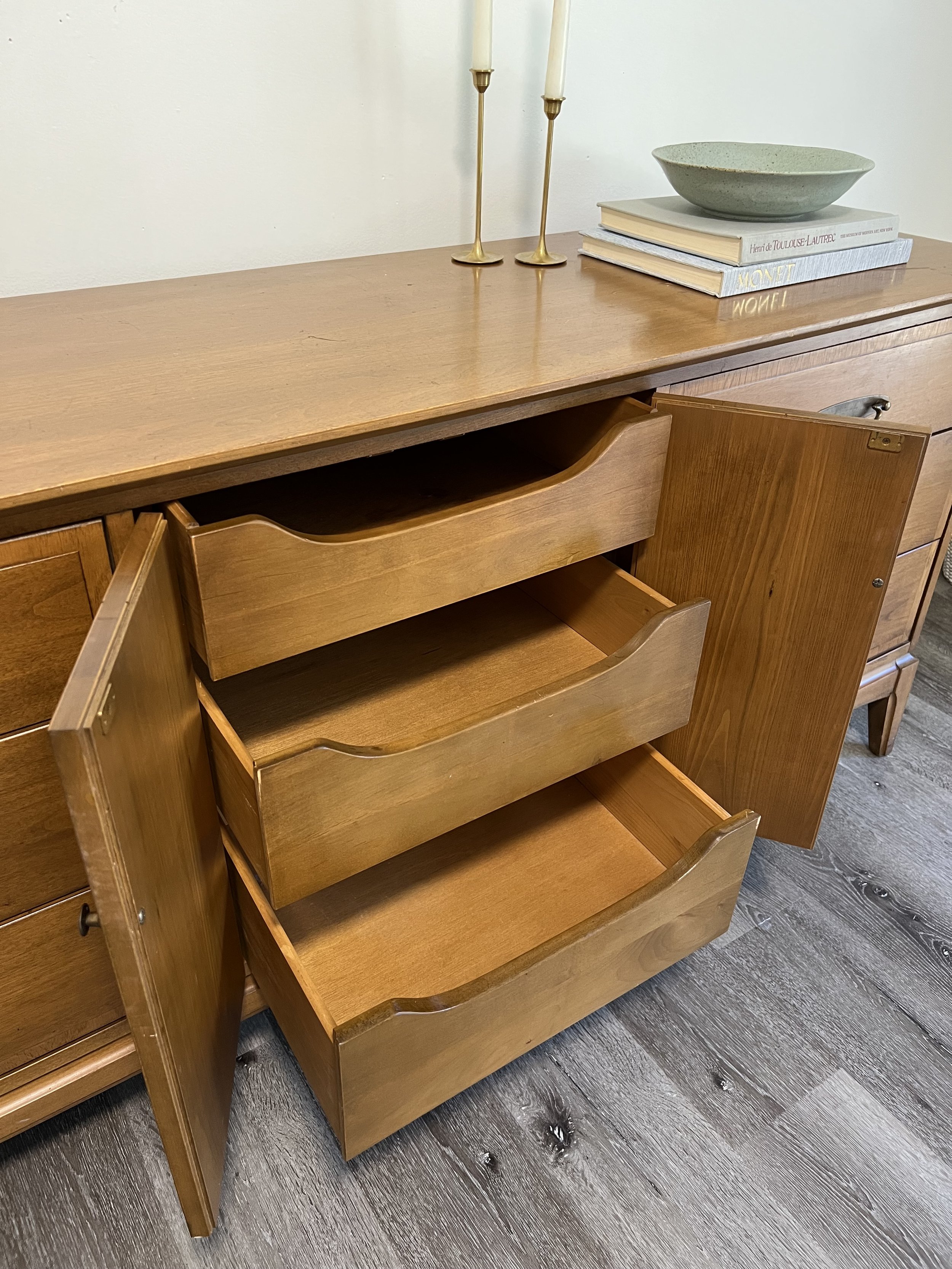 A wooden sideboard with three open drawers, and a matching handle on the closed door. On top, there are two gold candlesticks with white candles, a small stack of books, and a shallow green bowl.