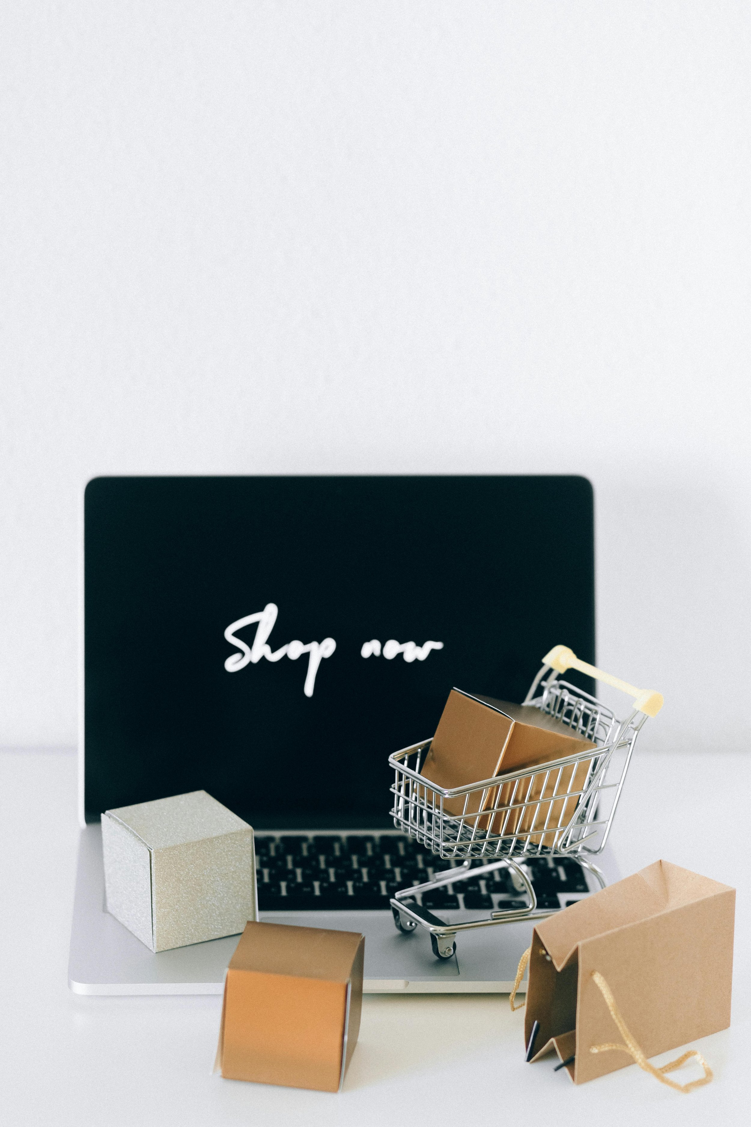Laptop with a shopping cart filled with boxes, surrounded by small wrapped boxes and shopping bags, on a white surface with a black screen displaying 'Shop now' in white text