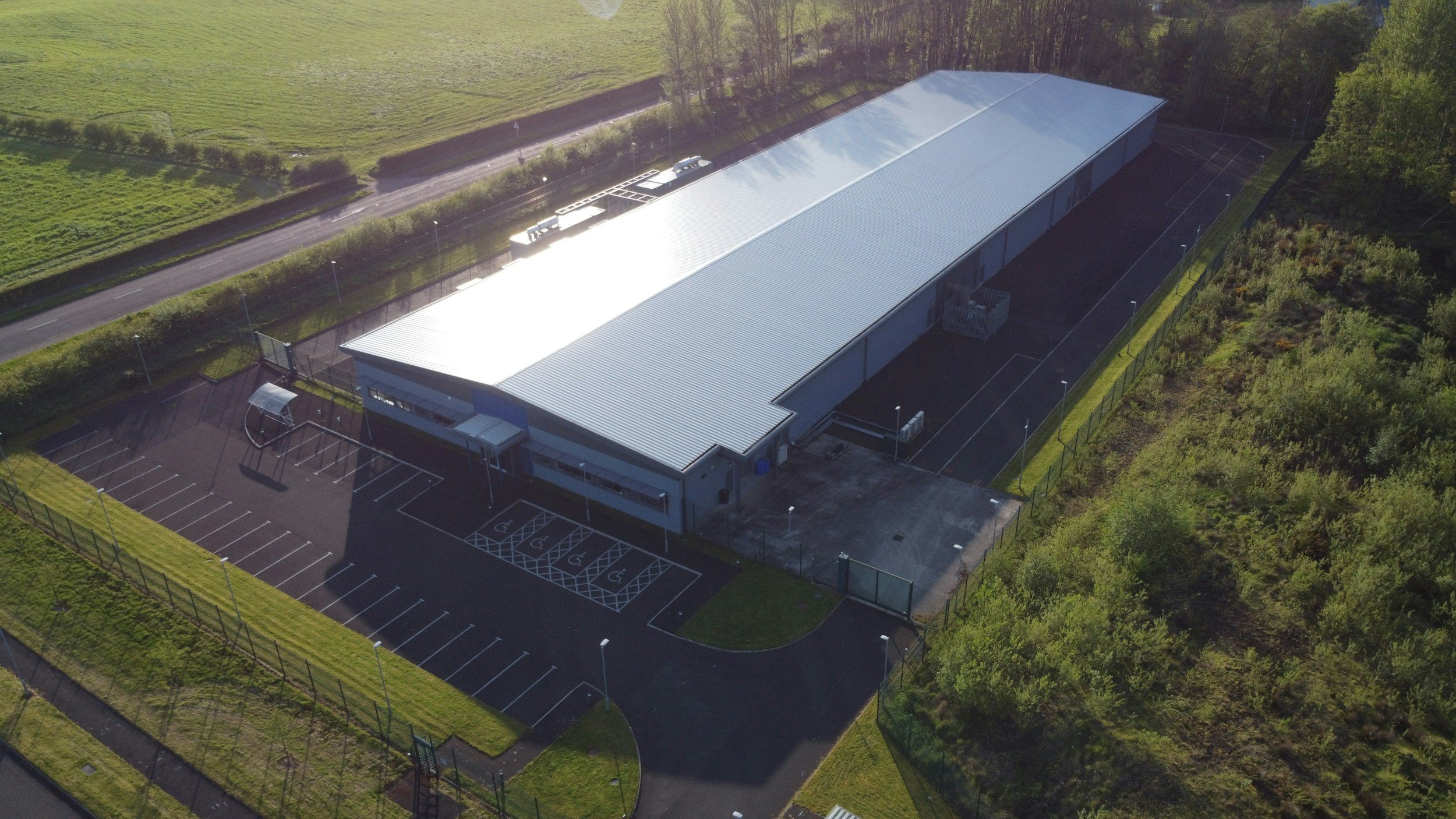Aerial view of a large, modern, silver-roofed sports facility with parking spaces, including accessible parking, surrounded by greenery and trees.