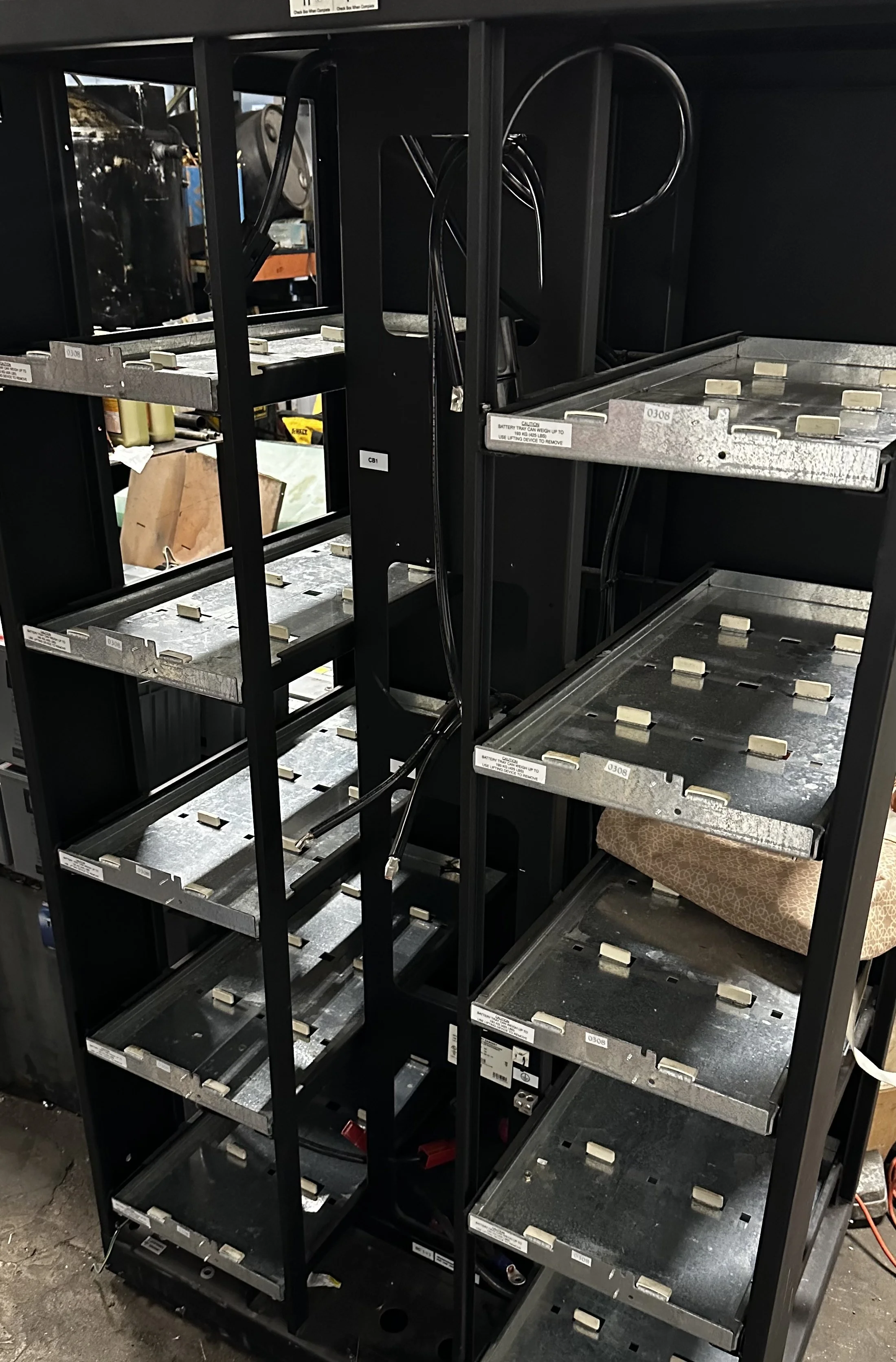 Empty black metal shelving unit with some attached wires, located in a storage or warehouse environment.
