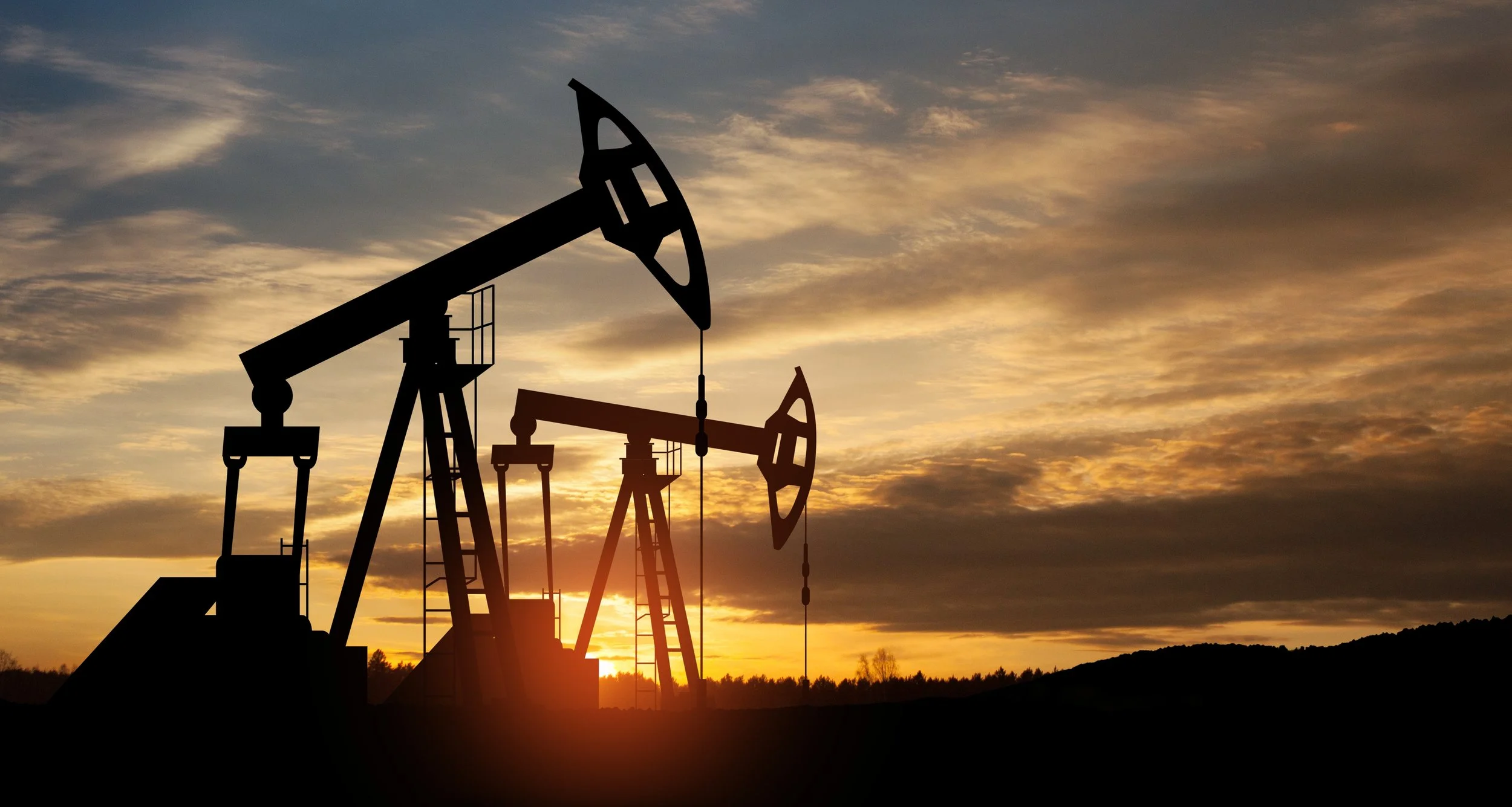 Silhouettes of oil pump jacks against a sunset sky with clouds and a distant treeline.