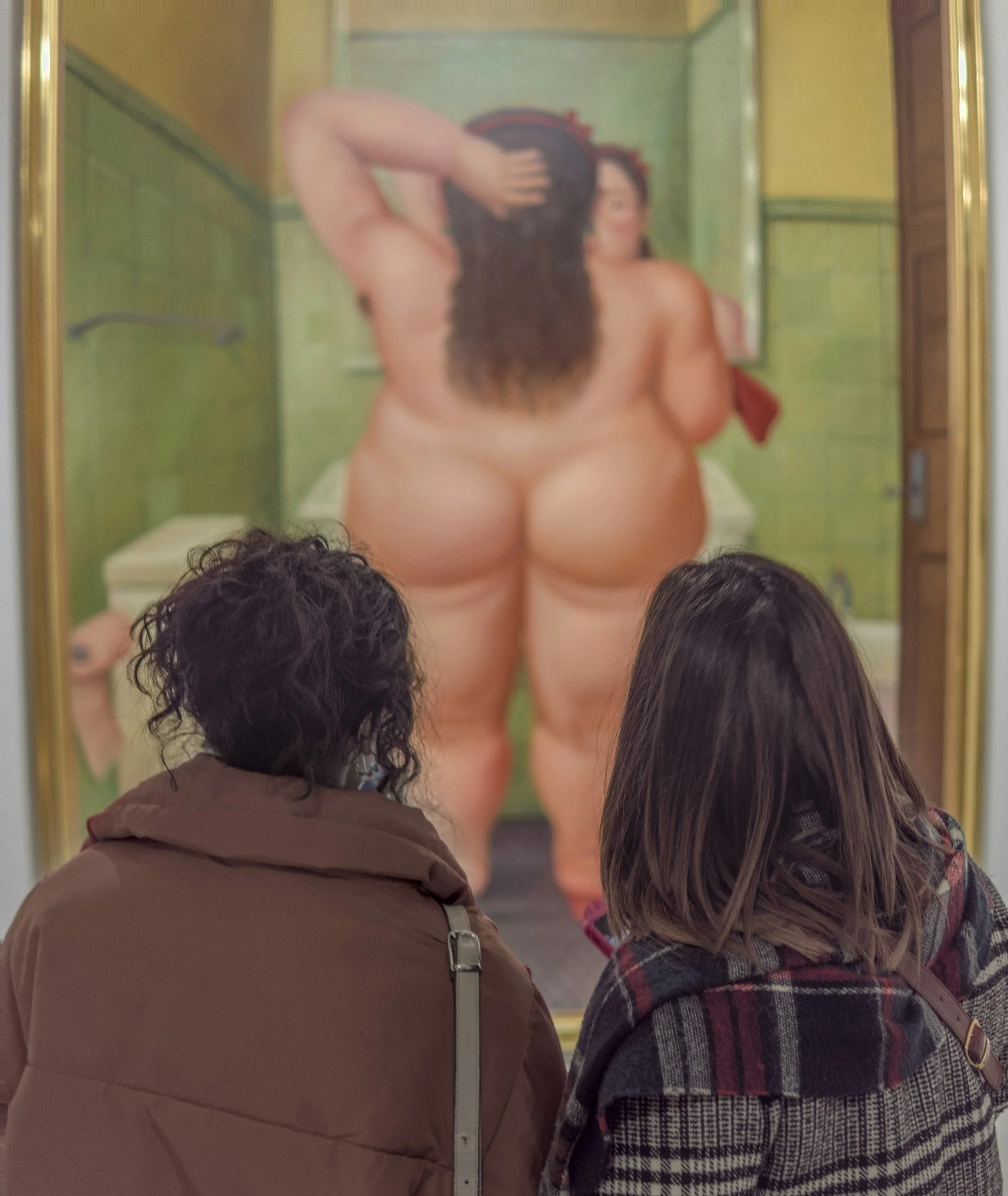 Two women are observing a large painting of a nude woman with her back facing the viewer at an art gallery.
