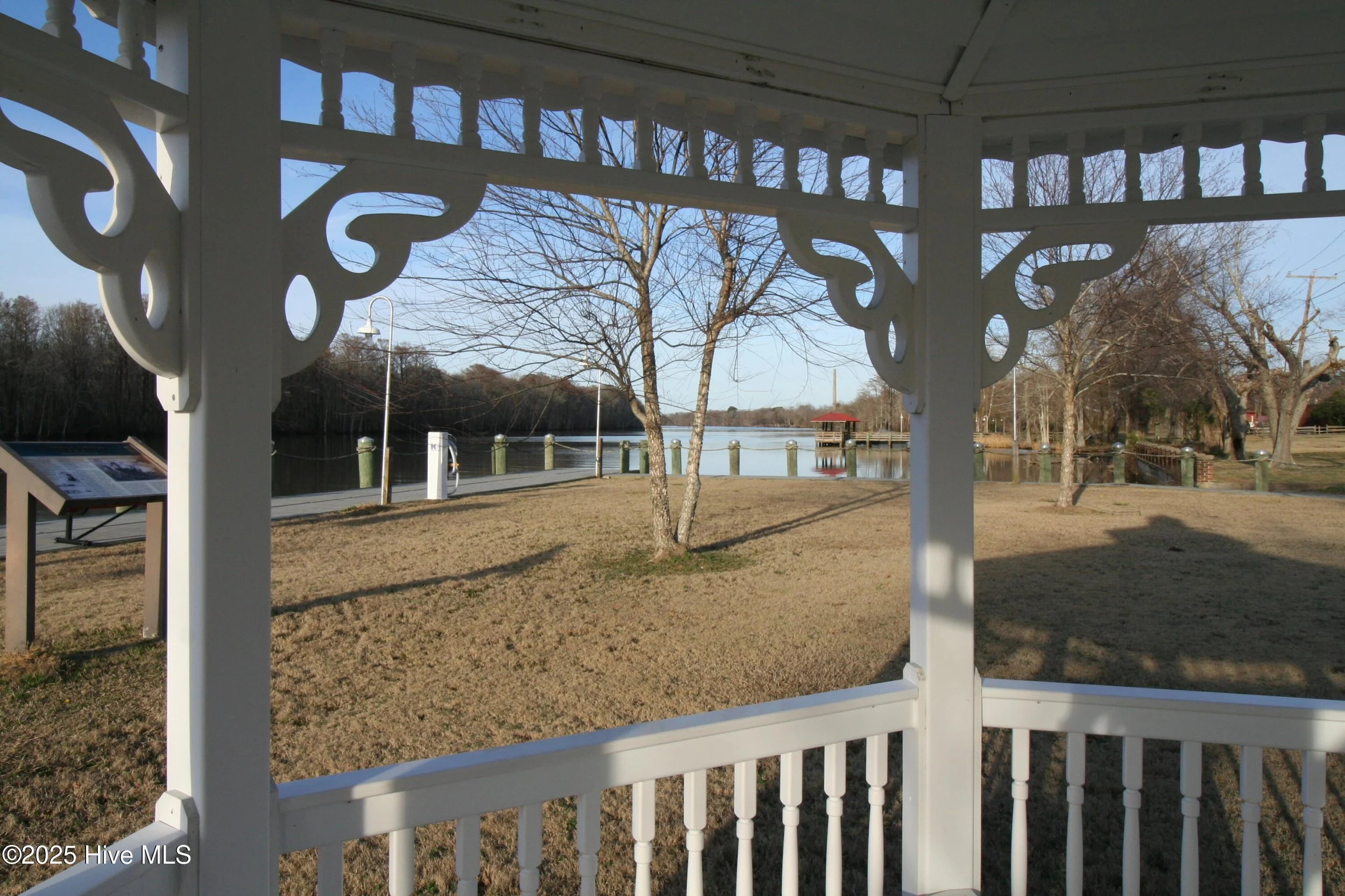View of a park with a lake, trees, and a dock seen through the white lattice and railing of a gazebo.