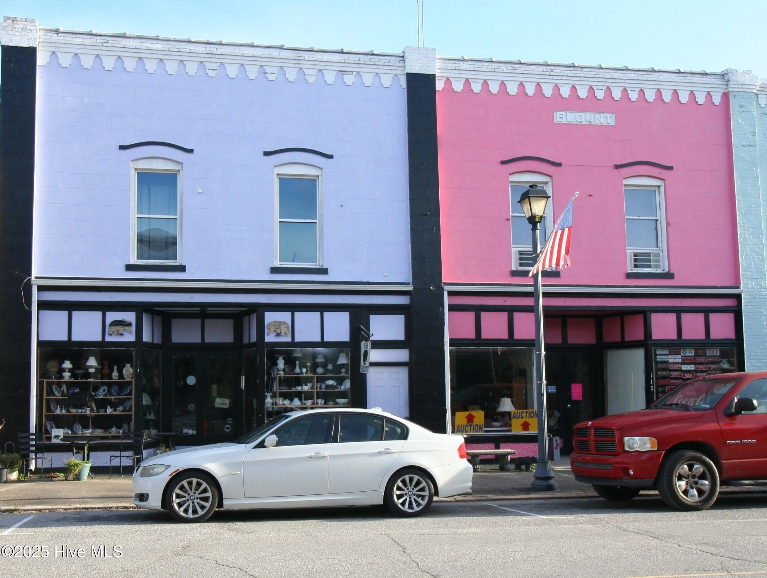 Two colorful buildings with painted faces and black eyebrows, one painted lavender and the other pink. The buildings have two floors each, with storefronts on the ground level displaying items.