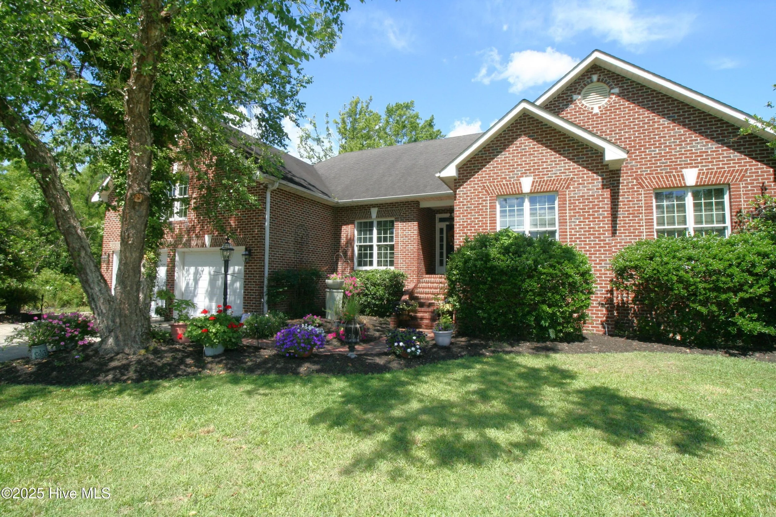 Red brick house with white window frames, front door, and landscaped front yard with bushes, flowers, and a tree, under a partly cloudy sky.