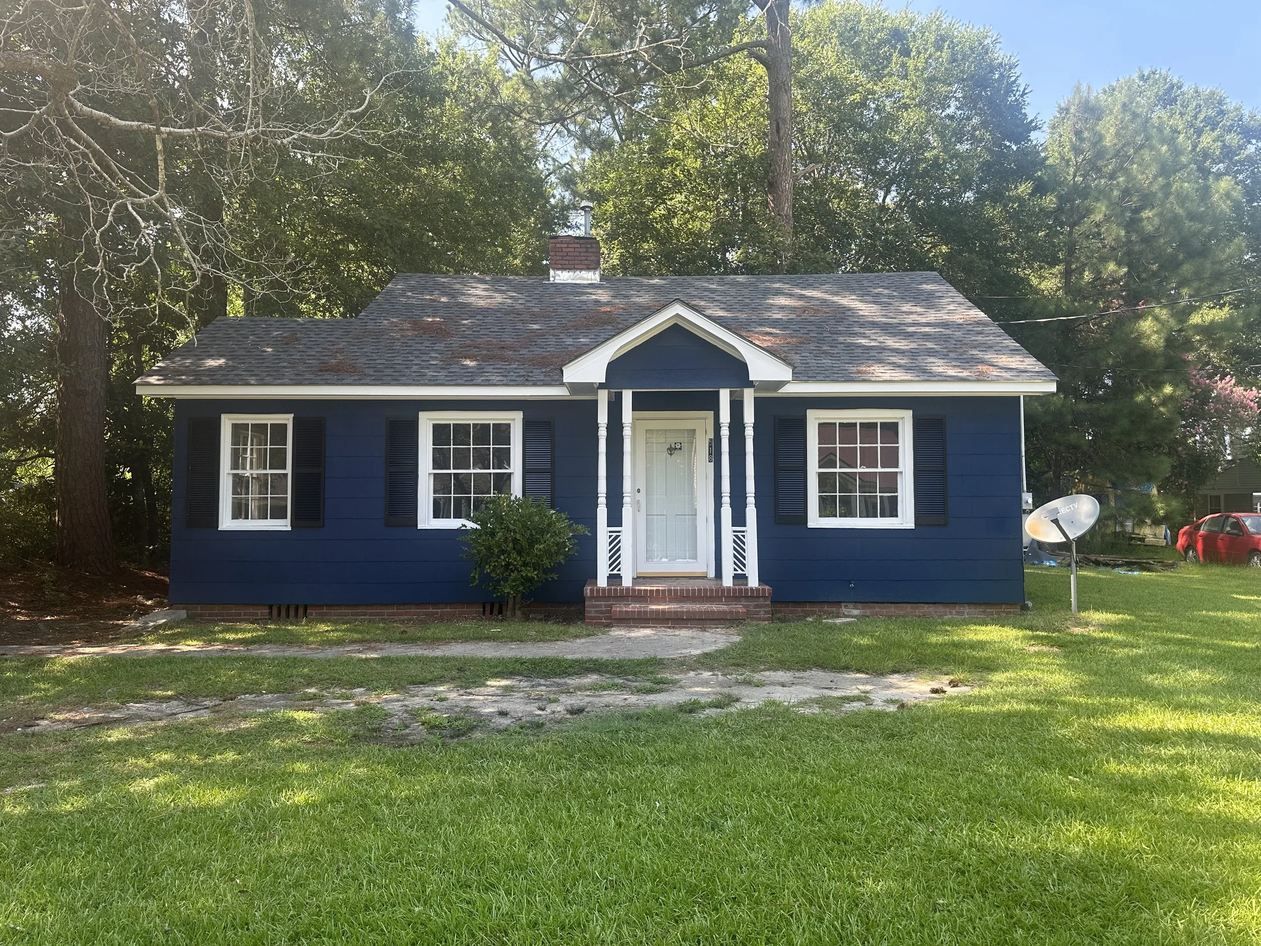 A small, charming house painted in navy blue with white trim, black shutters, and a brick front porch. The house has two windows on each side of the front door, which is centered and accessed by a small set of brick steps. There is a small bush in front of the house and a lawn with a driveway leading up to it. Tall trees surround the house, and there is a satellite dish on the right side.