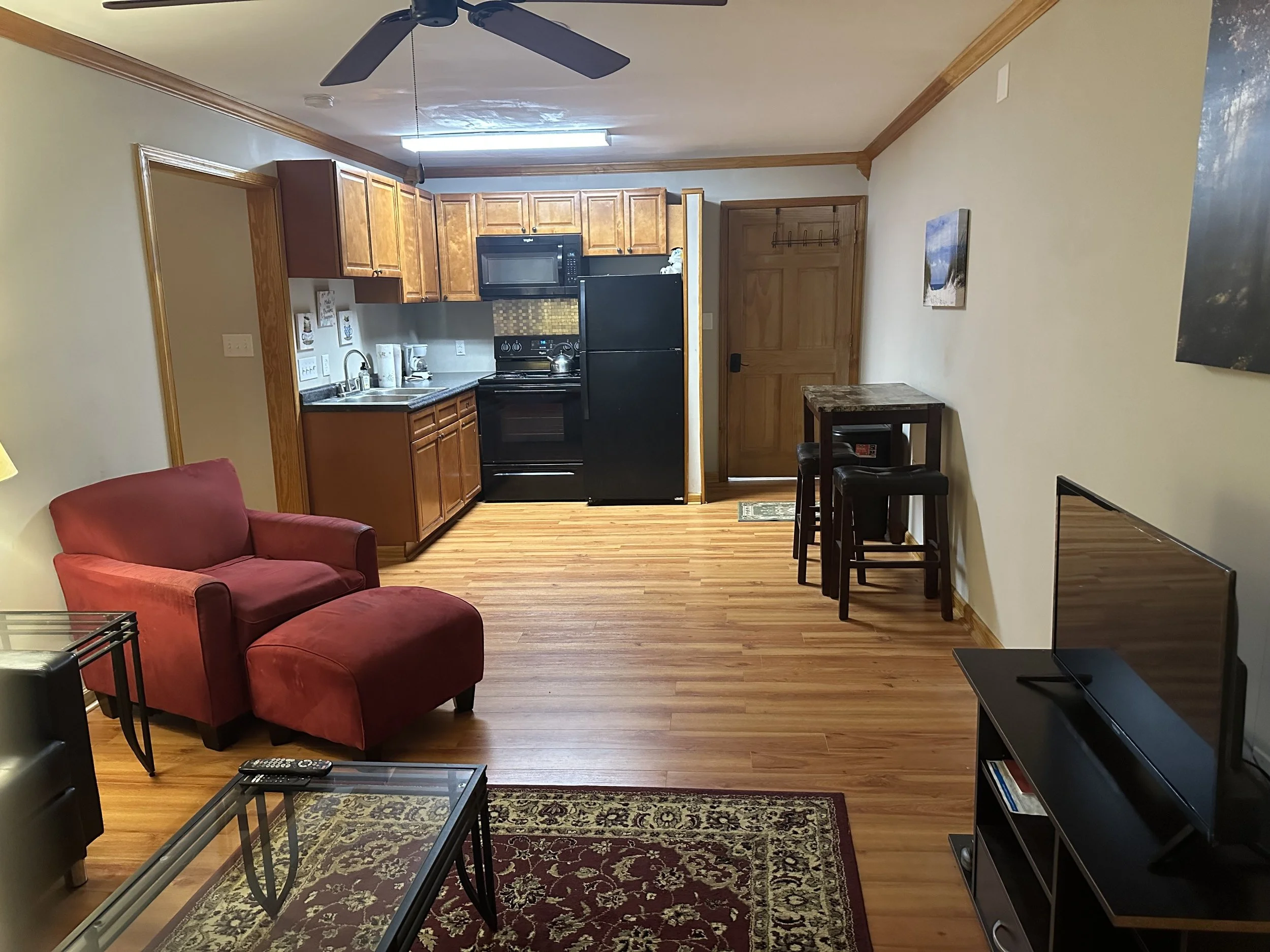 Living room and kitchen area with hardwood floors, red armchair with matching ottoman, glass coffee table, television on stand, kitchenette with black refrigerator, stove, microwave, and wooden cabinets, small high table with three bar stools, ceiling fan, and wall art.