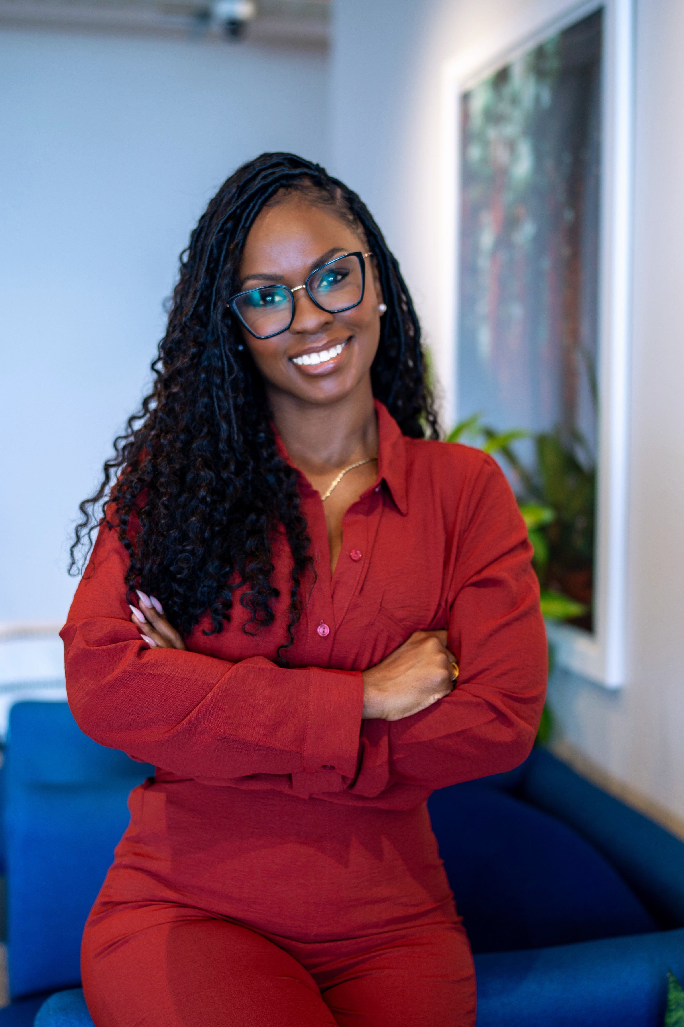 Portrait of a woman with long black hair, wearing glasses, a navy blazer, and a black top, standing against a plain gray background.