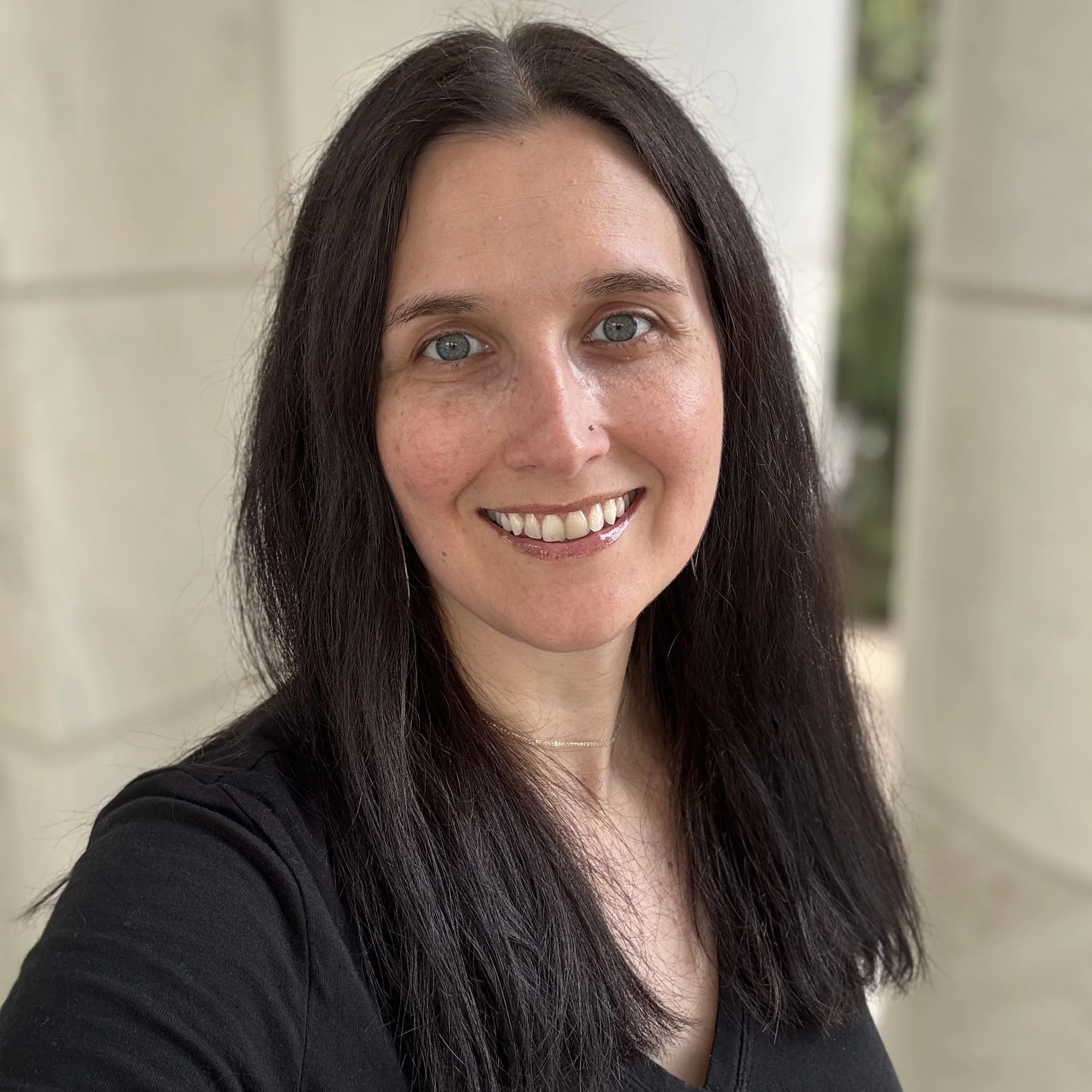 A woman with long dark hair, blue eyes, and a bright smile standing outdoors in front of a beige column and greenery.