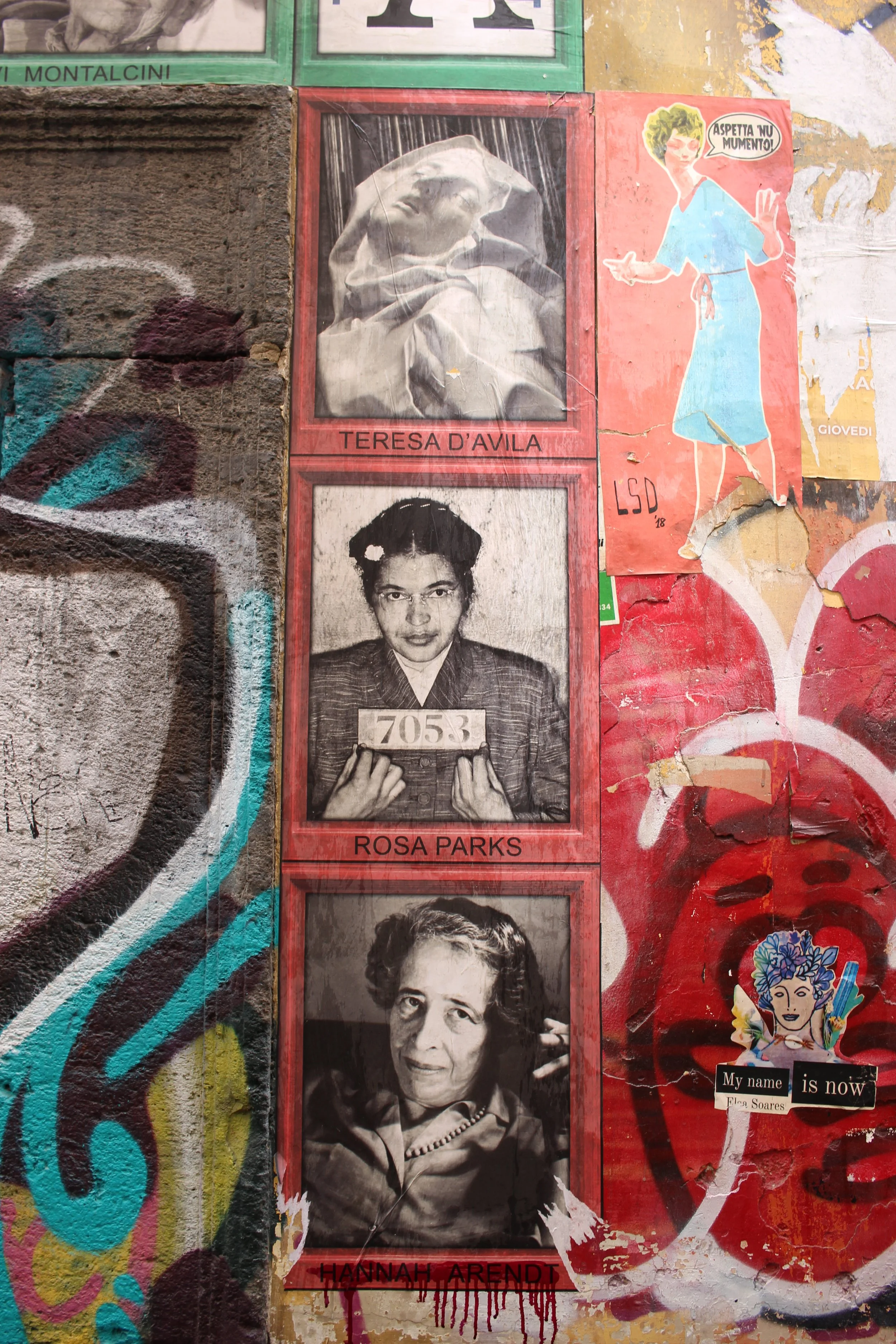 Graffiti und Street-Art-Poster mit drei schwarz-weißen Porträts, darunter Teresa D'Avila, Rosa Parks und Hannah Arendt, auf einer Wand mit bunten Graffiti. Außerdem gibt es eine Illustration eines Mädchens mit einem Sprechblasen-Text.