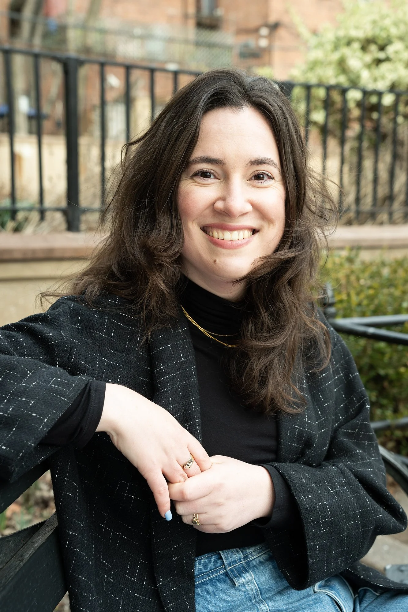 A woman with long brown hair, smiling, wearing a black turtleneck, black blazer with a checkered pattern, and jeans, sitting on a park bench outdoors.