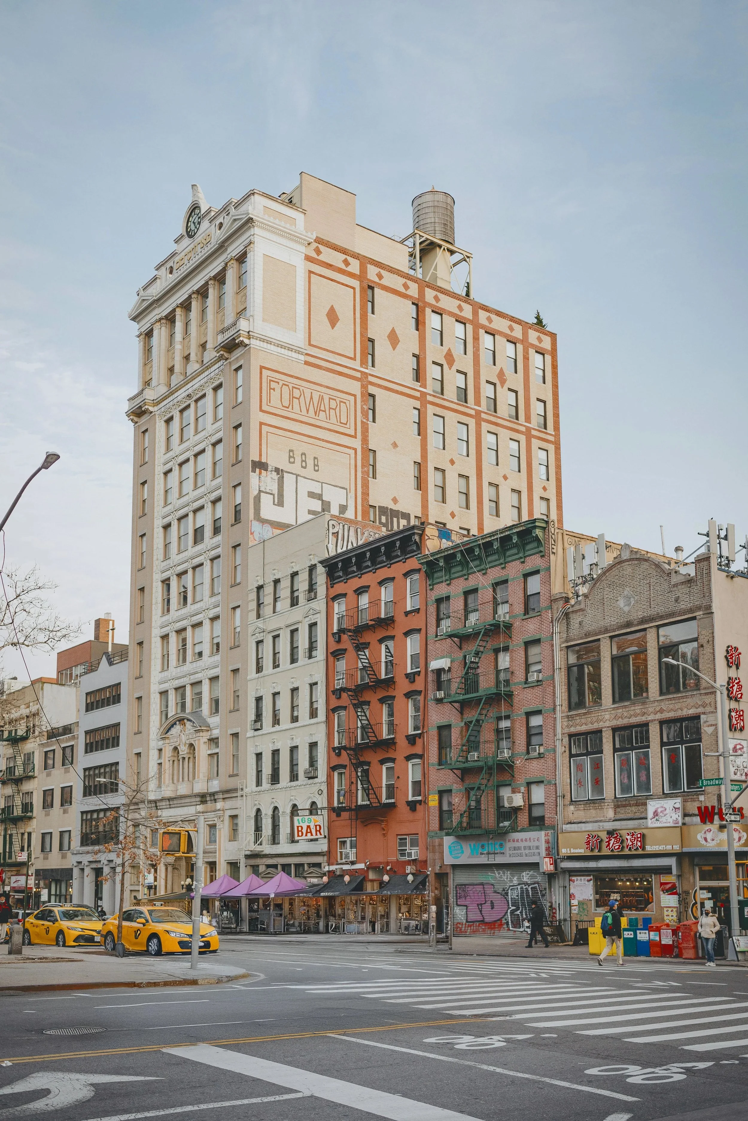 City street scene featuring a tall beige building with the words 'Forward' and 'Jet' graffiti, smaller colorful buildings with fire escapes, yellow taxis, pedestrians, sidewalk cafes with purple umbrellas, and storefront signs.
