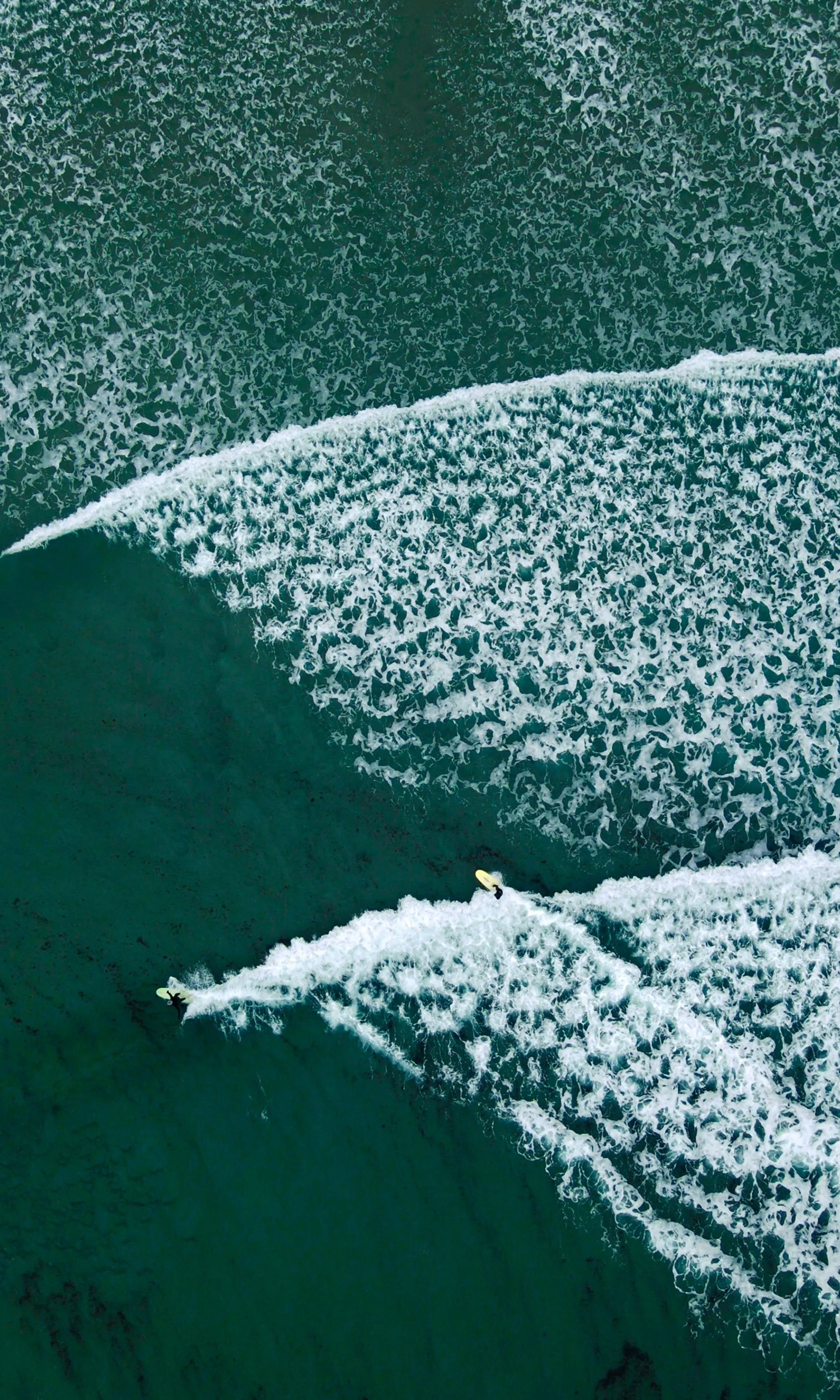 Two surfers riding the waves in the ocean, leaving white trails behind them on the deep blue water.