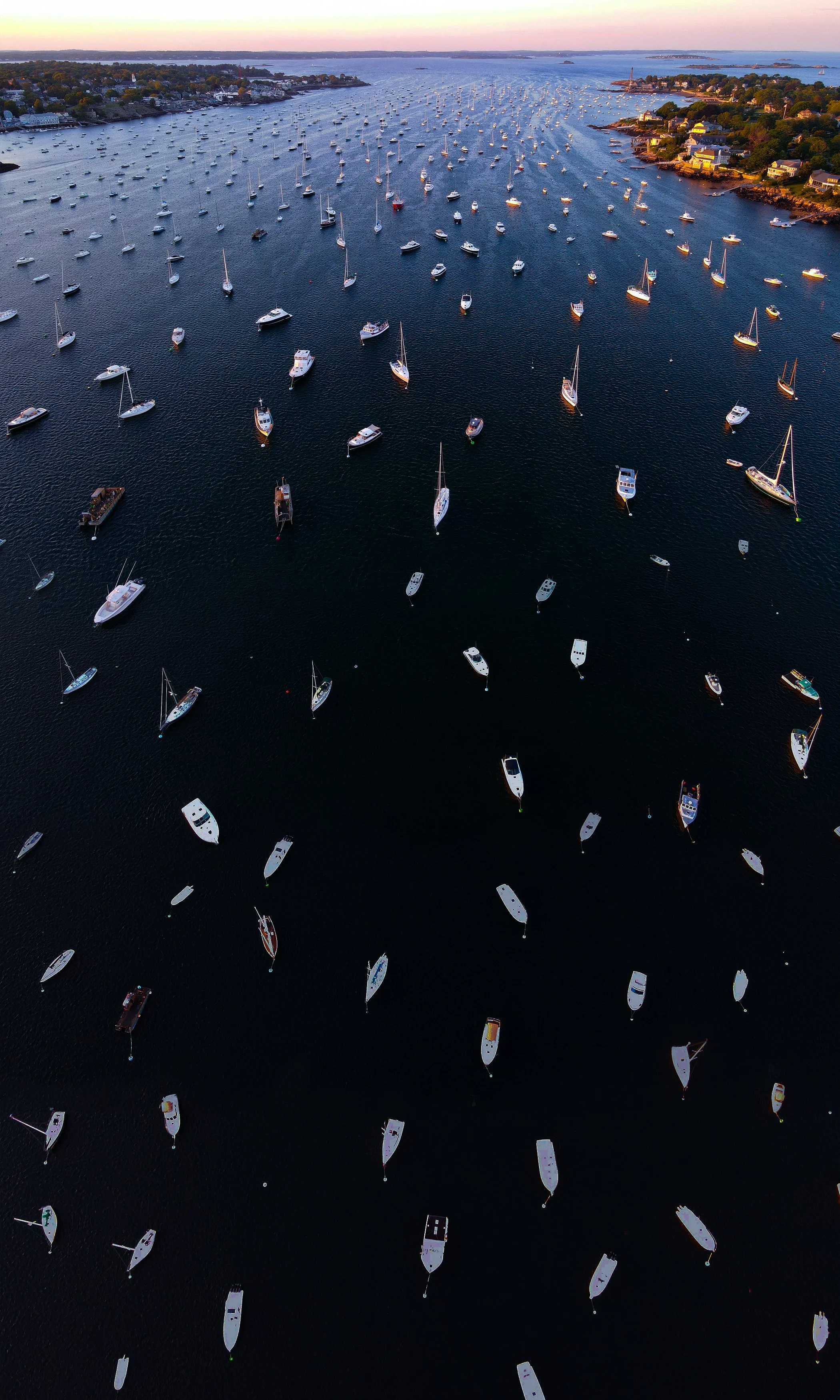 Aerial view of a harbor filled with numerous sailboats and yachts at sunset, with houses along the shoreline and distant islands.