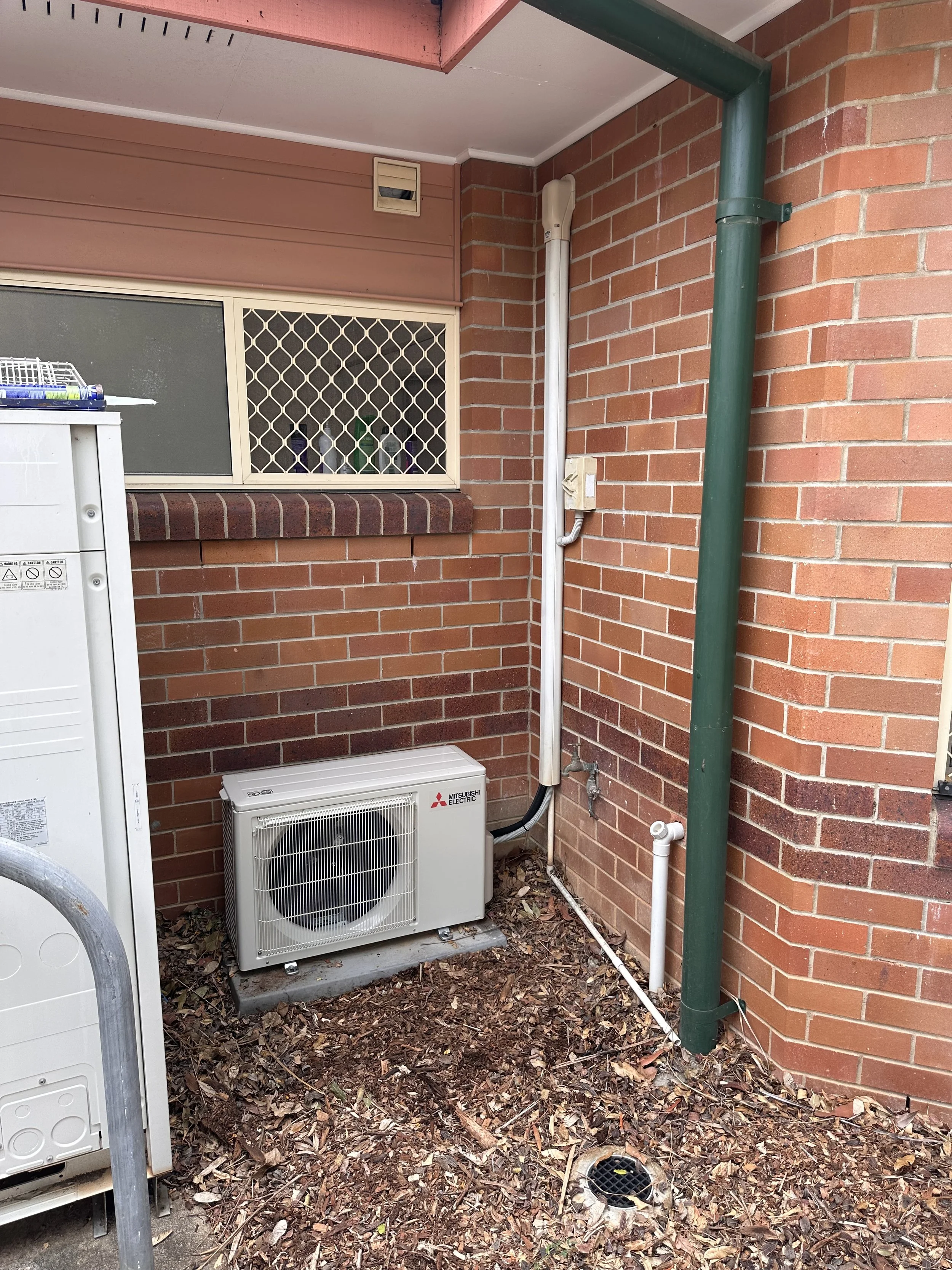 An outdoor corner with brick walls, a white Mitsubishi Electric air conditioning unit on a concrete slab, two drainage pipes, a window with a security grille, and a vent near the ceiling. The ground is covered with dry leaves.