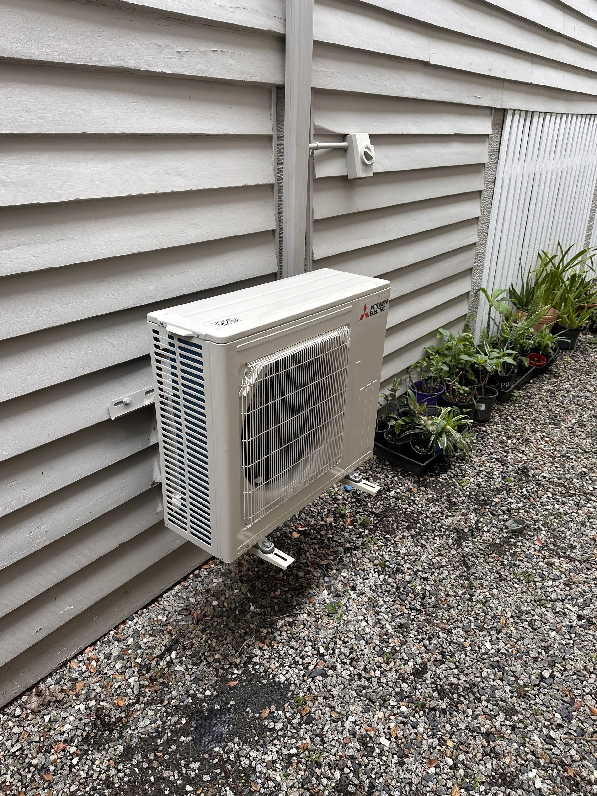 A white Mitsubishi Electric outdoor air conditioning unit mounted on a gravel ground next to a house with white horizontal siding. There are potted plants with green foliage along the house wall.