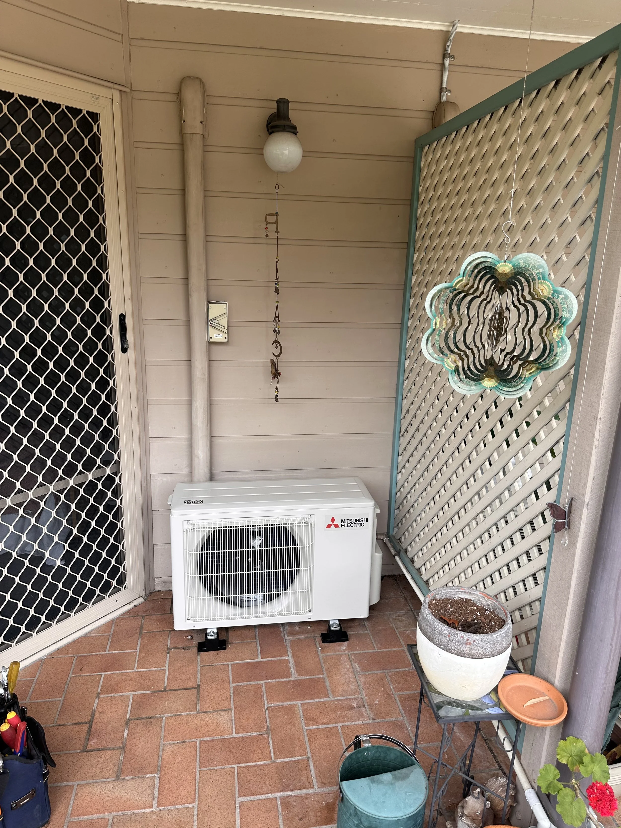 An outdoor patio area with a beige air conditioning unit labeled Mitsubishi Electric, a patterned hanging decoration, a terracotta planter with dried soil, and various gardening supplies and plants. The area has a lattice panel, a security door, and 